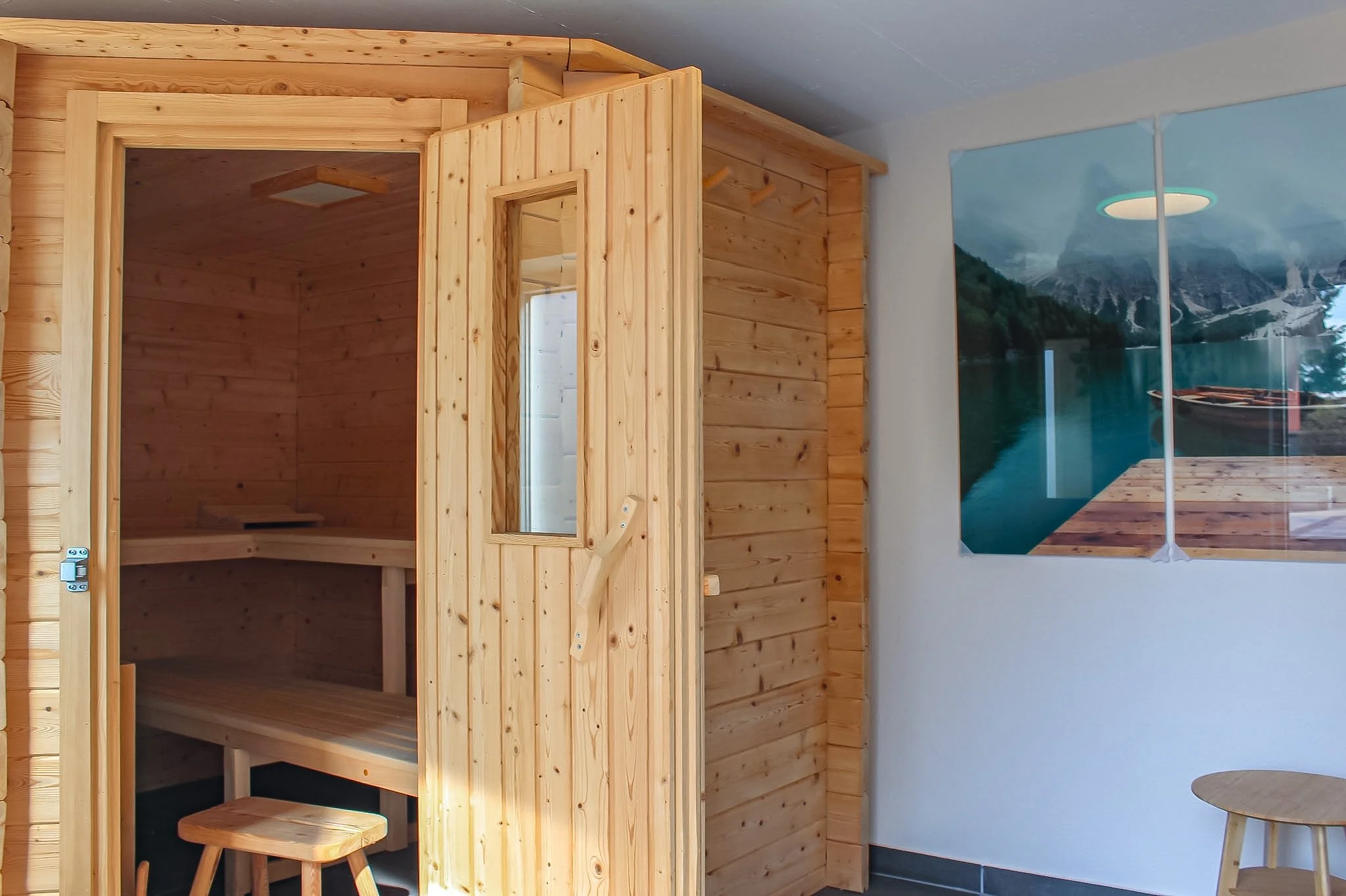 Innenaufnahme eines Raumes mit Holz-Inneneinrichtungen, darunter ein Holzofen oder Sauna, und ein Fenster mit Blick auf eine Berglandschaft mit glitzerndem See im Hintergrund.