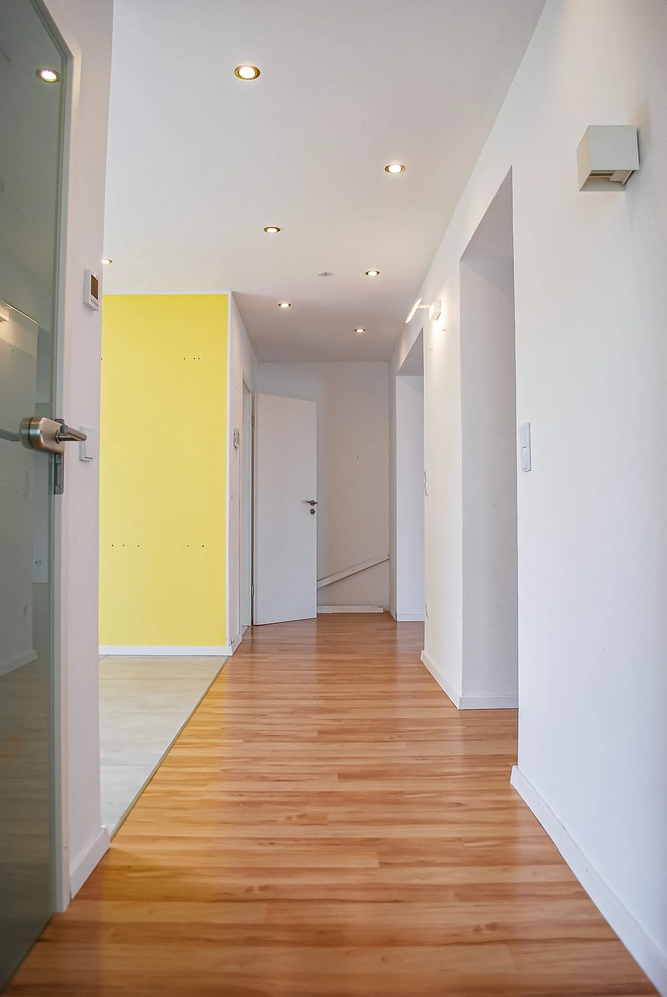 Moderner Flur mit Holzboden, weißen Wänden und Decke, mehreren Deckenspots, einer gelben Wand im Hintergrund, einer Tür, die nach rechts führt, und einer Glaswand auf der linken Seite.