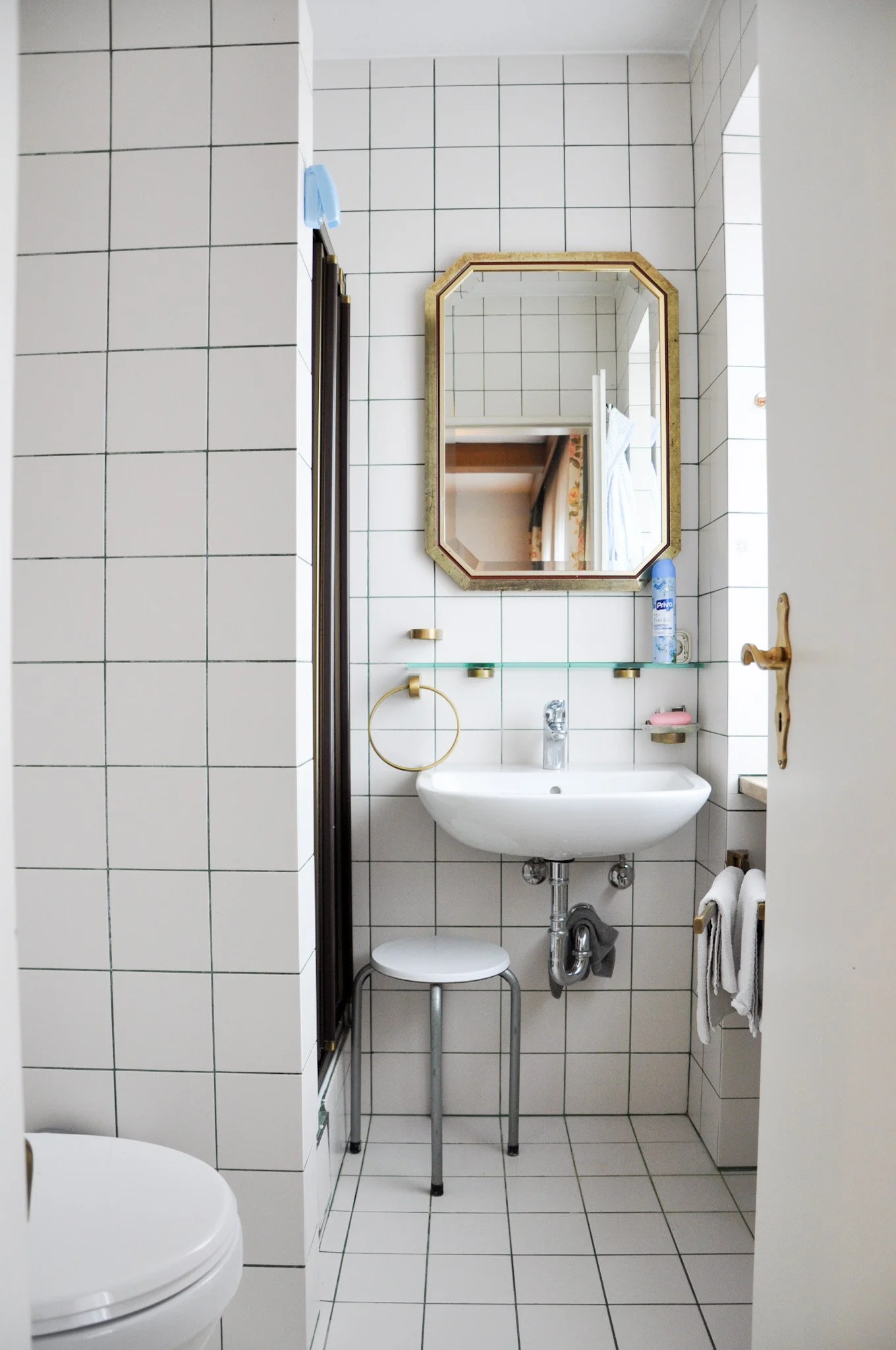 Ein kleines Badezimmer mit weißen Wandfliesen, einem Wandspiegel, Waschbecken, einem Toilettenhocker, Handtüchern, Seifenschale, einem Handtuchhalter und Reinigungsmitteln.