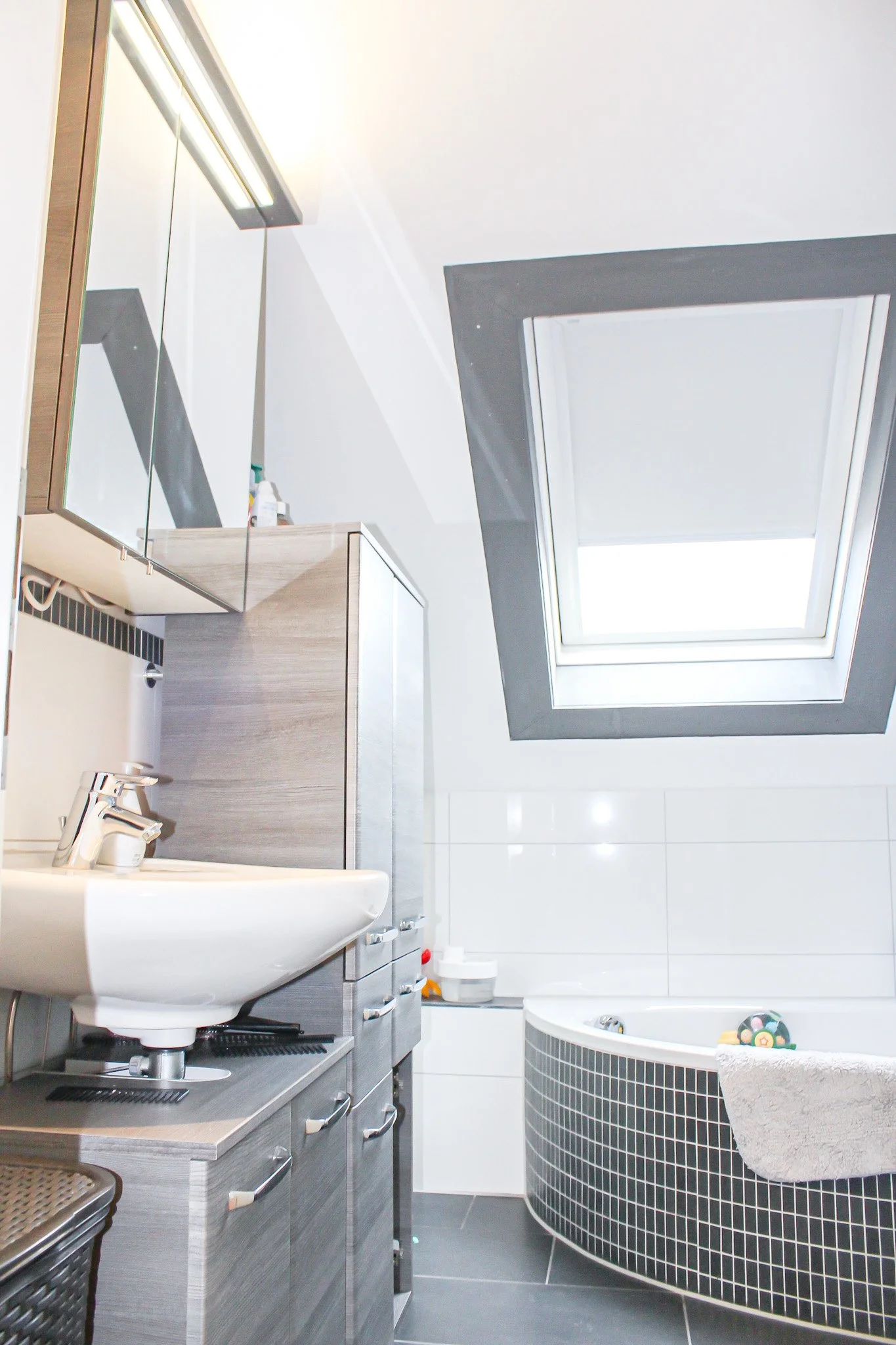 Ein modernes Badezimmer mit Waschbecken, dunklen Schränken, einem runden Wannenbad mit schwarzen Fliesen, einem großen Dachfenster, weißen Wänden und einer dunklen Decke.