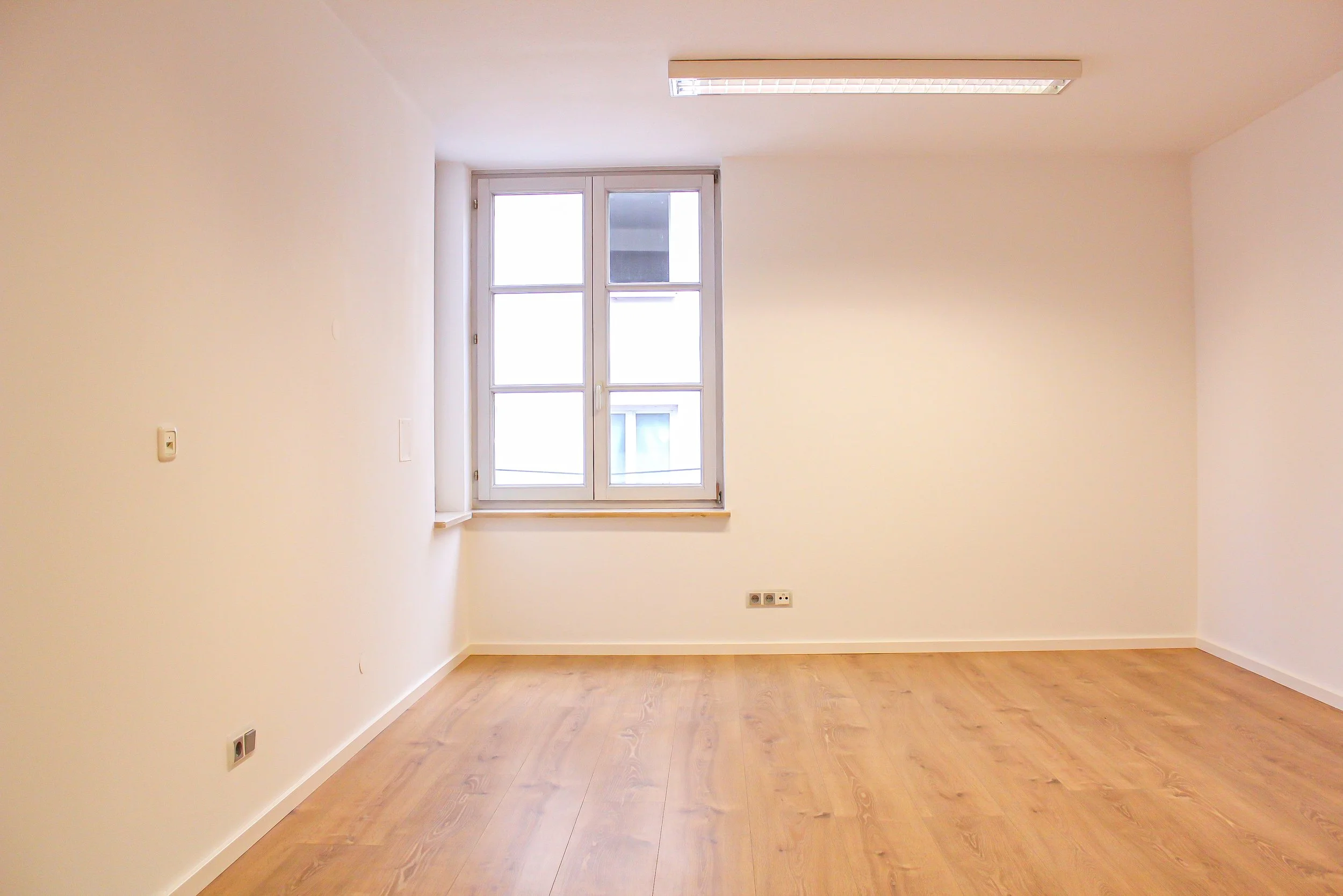 Leeres Zimmer mit großen Fenster, weißen Wänden und Holzfußboden.