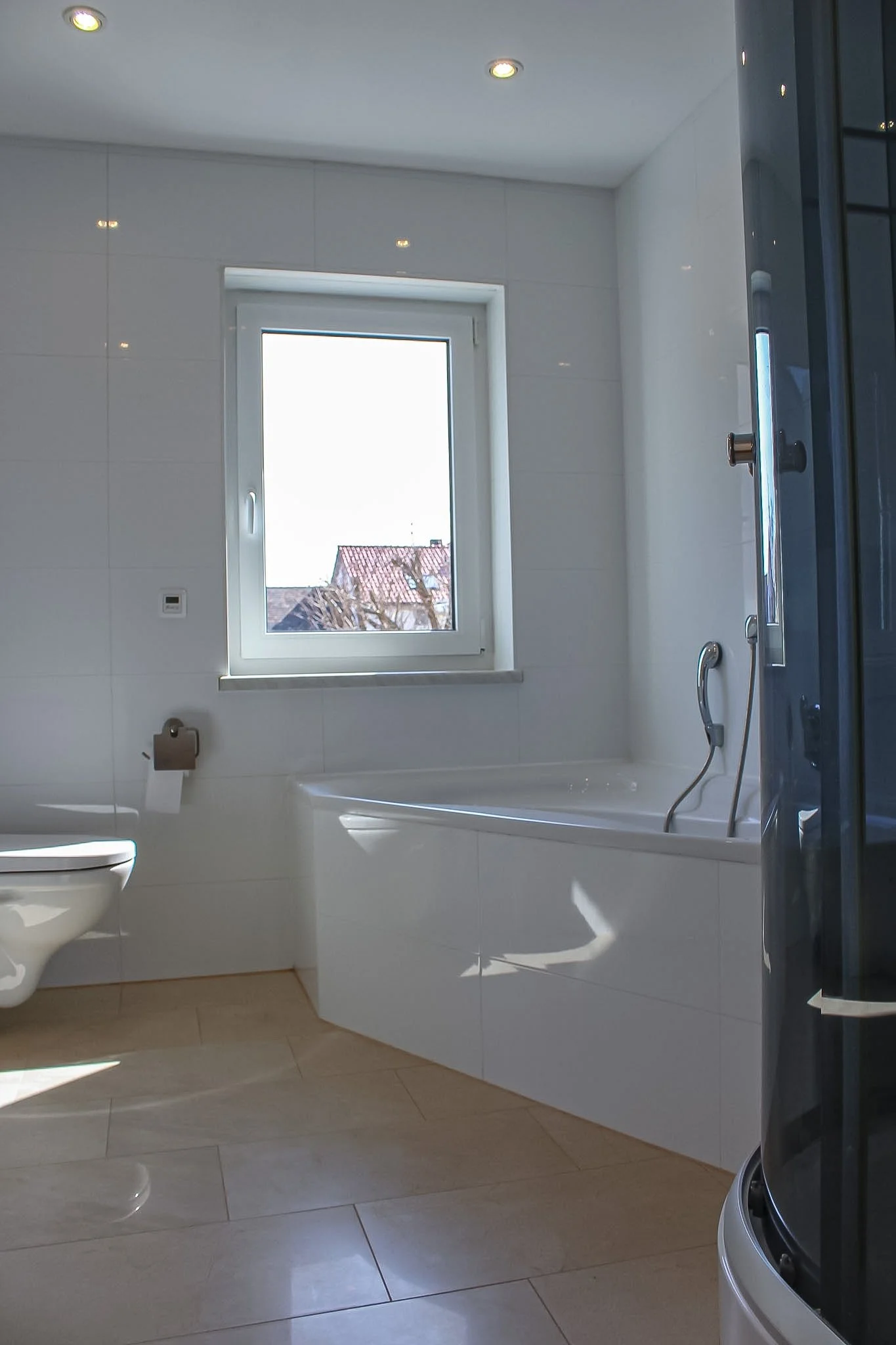 Ein Badezimmer mit weiß gefliesten Wänden, einer Badewanne, einem Fenster mit Blick auf Dächer und einem Duschbereich mit Glasduschkabine.