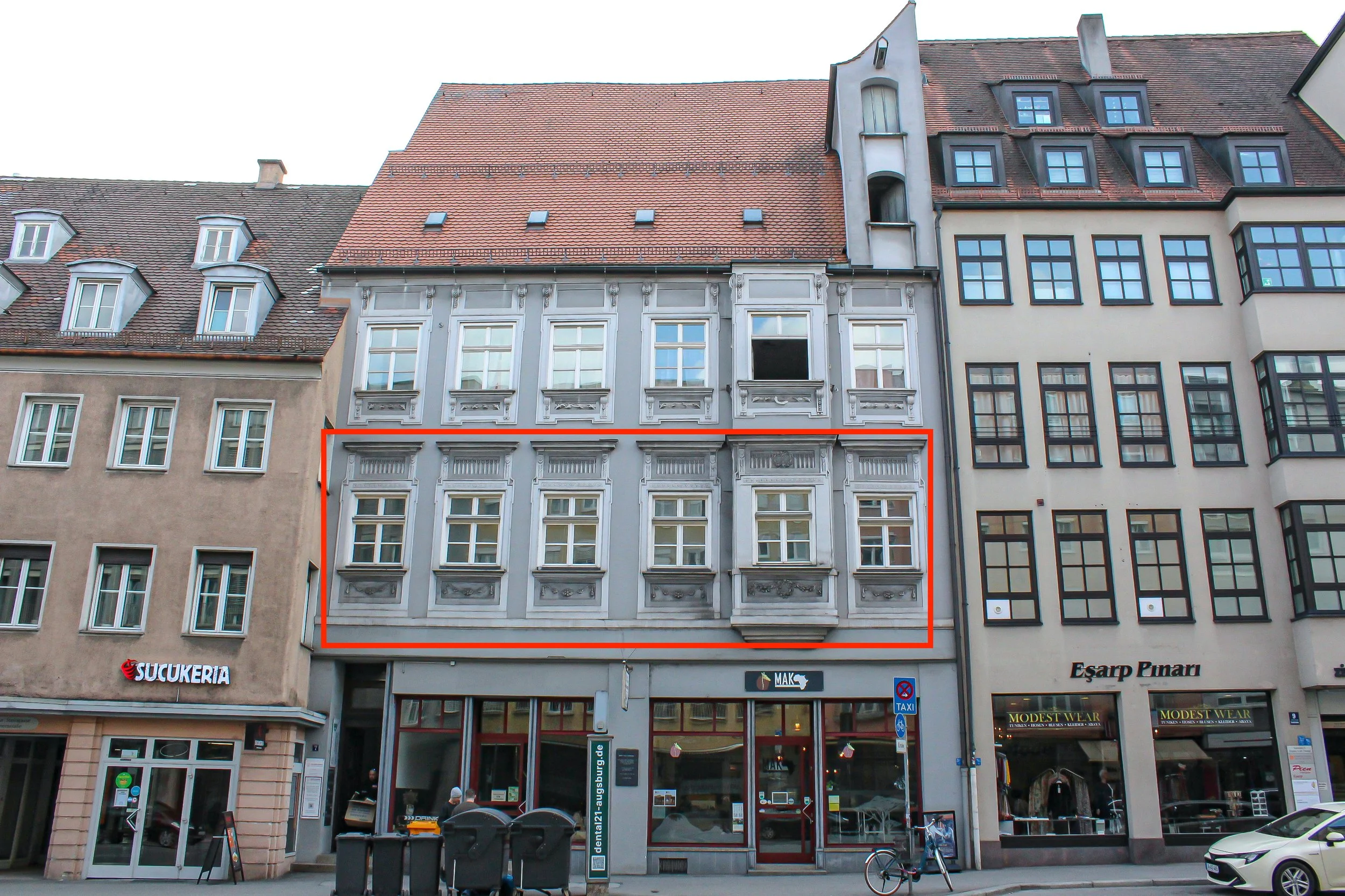 Mehrstöckiges Gebäude mit barocker Architektur an der Vorderseite, im unteren Bereich Geschäfte und Fahrräder, davor Mülltonnen, mehrere Fenster und ein Auftragsschneidegerät.