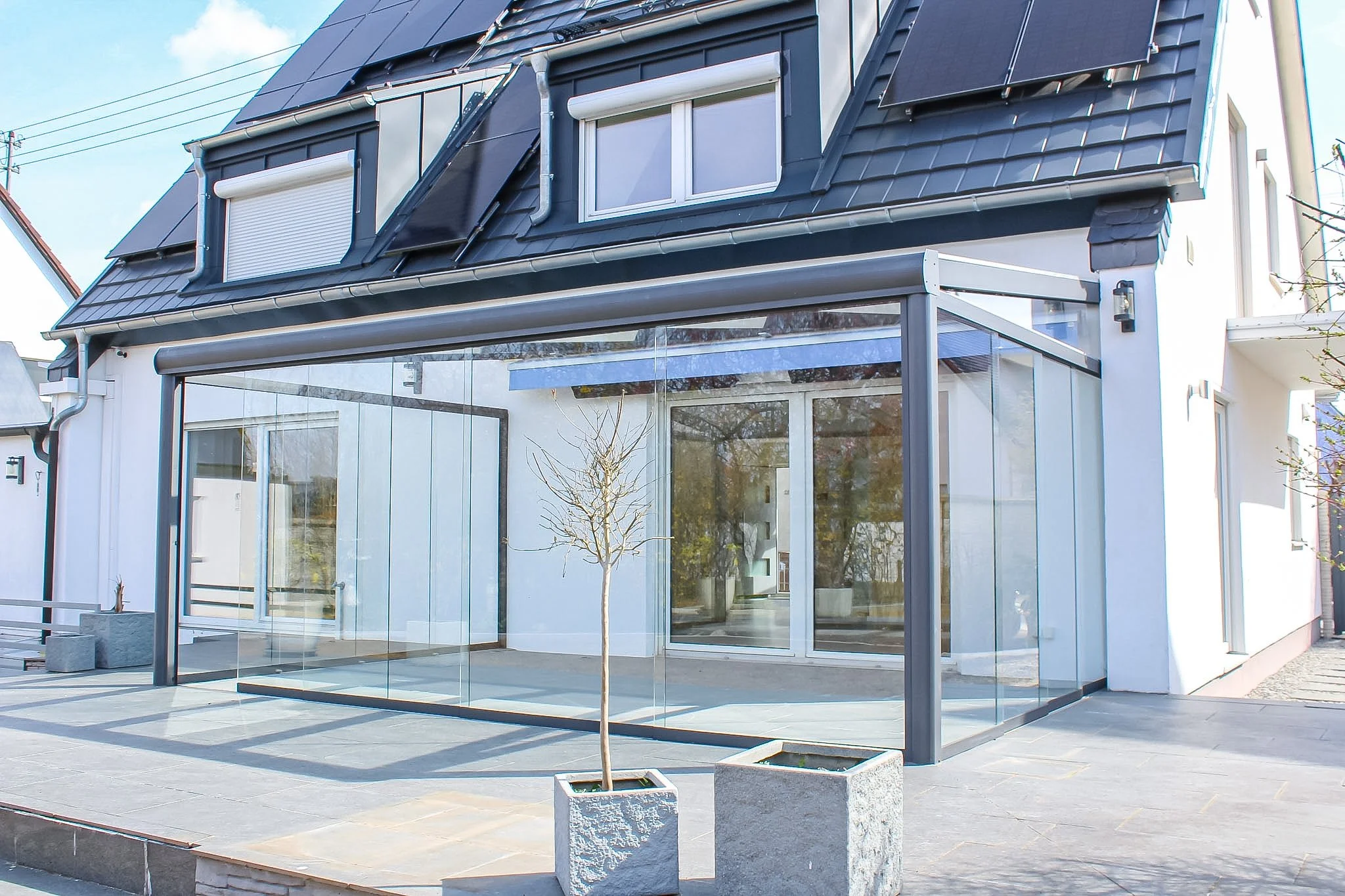 Moderne Hausfassade mit großen Glasfenstern, einem Wintergarten, einem kleinen Baum in einem Quadratgarten und einem gepflasterten Außenbereich.