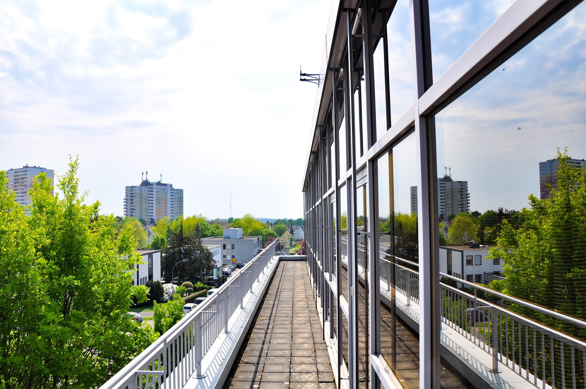 Blick von einem Balkon eines modernen Gebäudes mit großen Fenstern auf eine grüne Parkanlage und hochtecityfühlte Stadt mit mehreren Hochhäusern im Hintergrund.