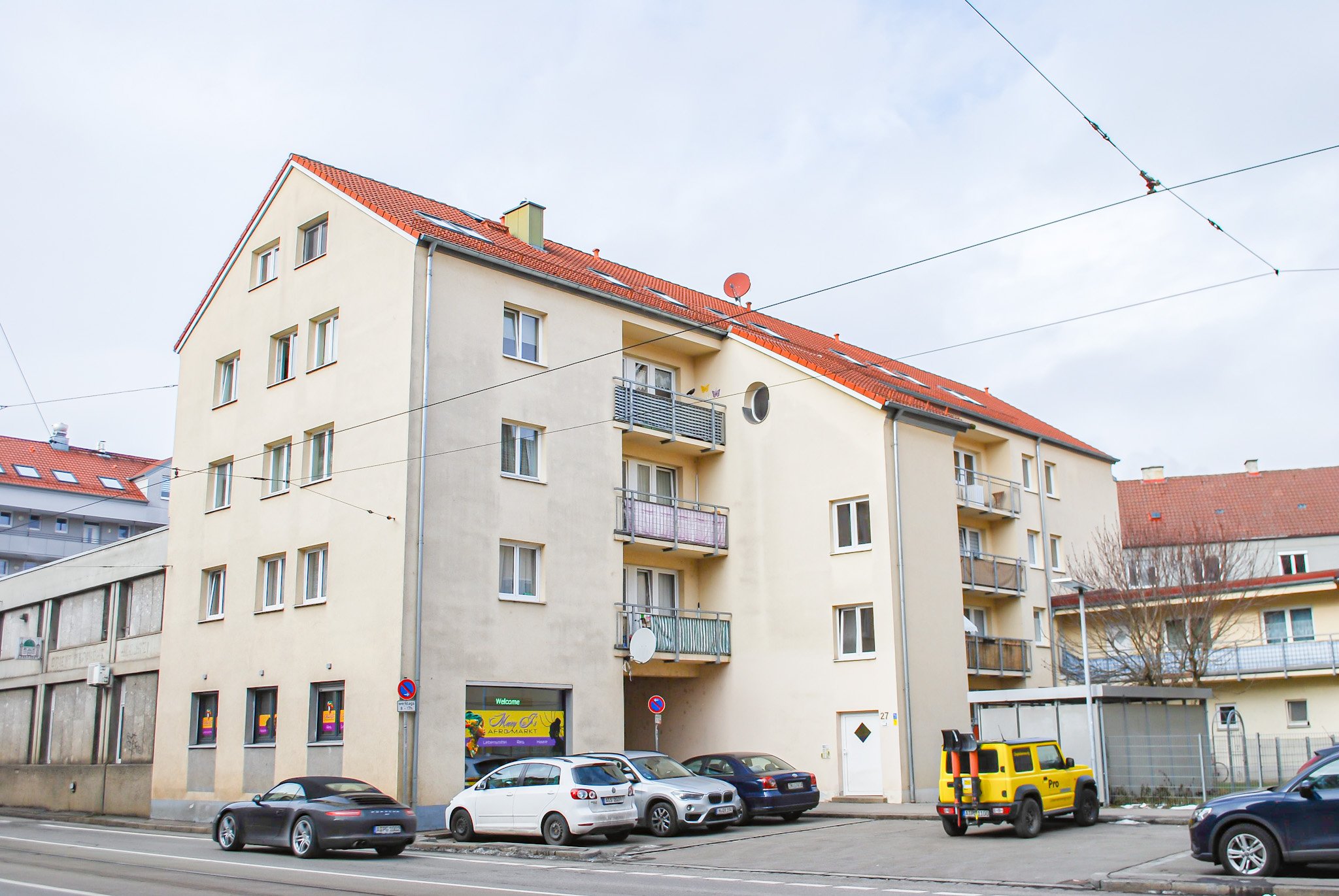 Mehrstöckiges Mehrfamilienhaus mit roten Ziegeldach, mehreren Balkonen, Parkplätzen und Straßenverkehr in einer städtischen Umgebung.