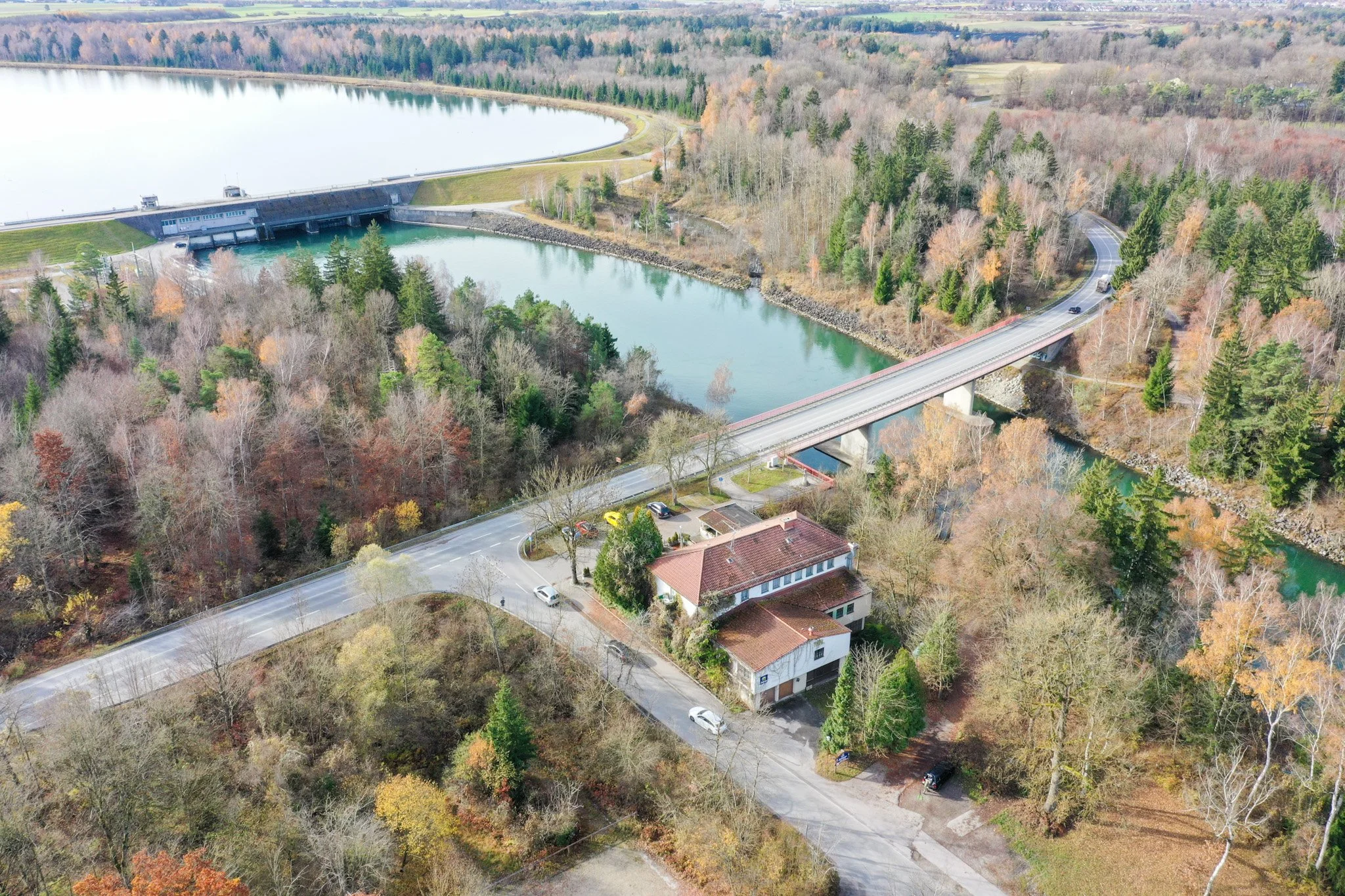 Luftaufnahme von einem Wasserreservoir, umgeben von Bäumen im Herbst, mit einer Brücke und kleinen Gebäuden im Vordergrund.