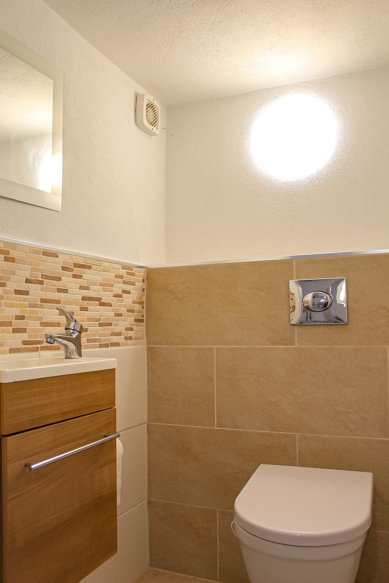 Ein Badezimmer mit einem Waschbecken, einer Holzschublade, einem rechteckigen Spiegel, einer braunen Wand mit Fliesen und einer weißen Toilette. Es gibt eine Wandheizung oder Abzug im oberen Bereich der Wand.