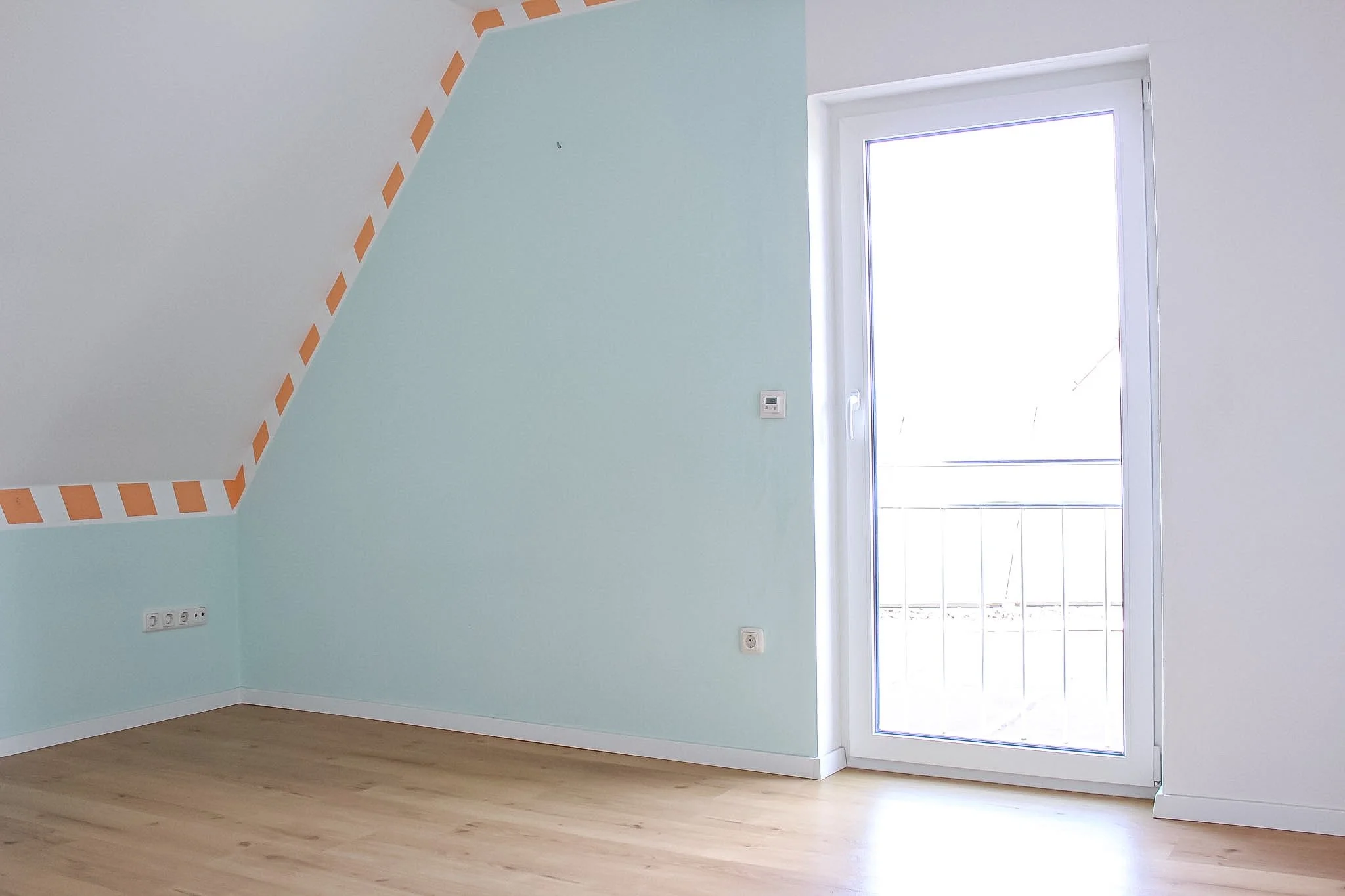 Ein leeres Zimmer mit hellblauen Wänden, weißen Rahmen, Holzboden und einem Balkon mit Glasfenster.