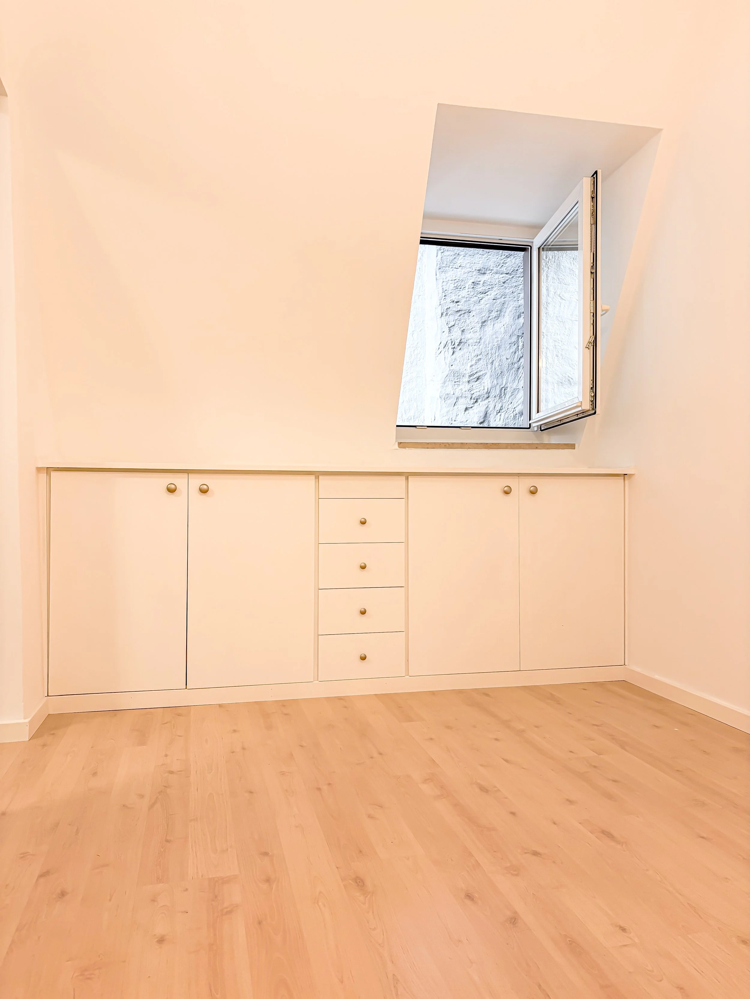 Ein weißer Einbauschrank mit mehreren Türen und Schubladen in einem Raum mit Holzfußboden. Ein offenes Fenster mit Blick auf eine weiße Mauer.