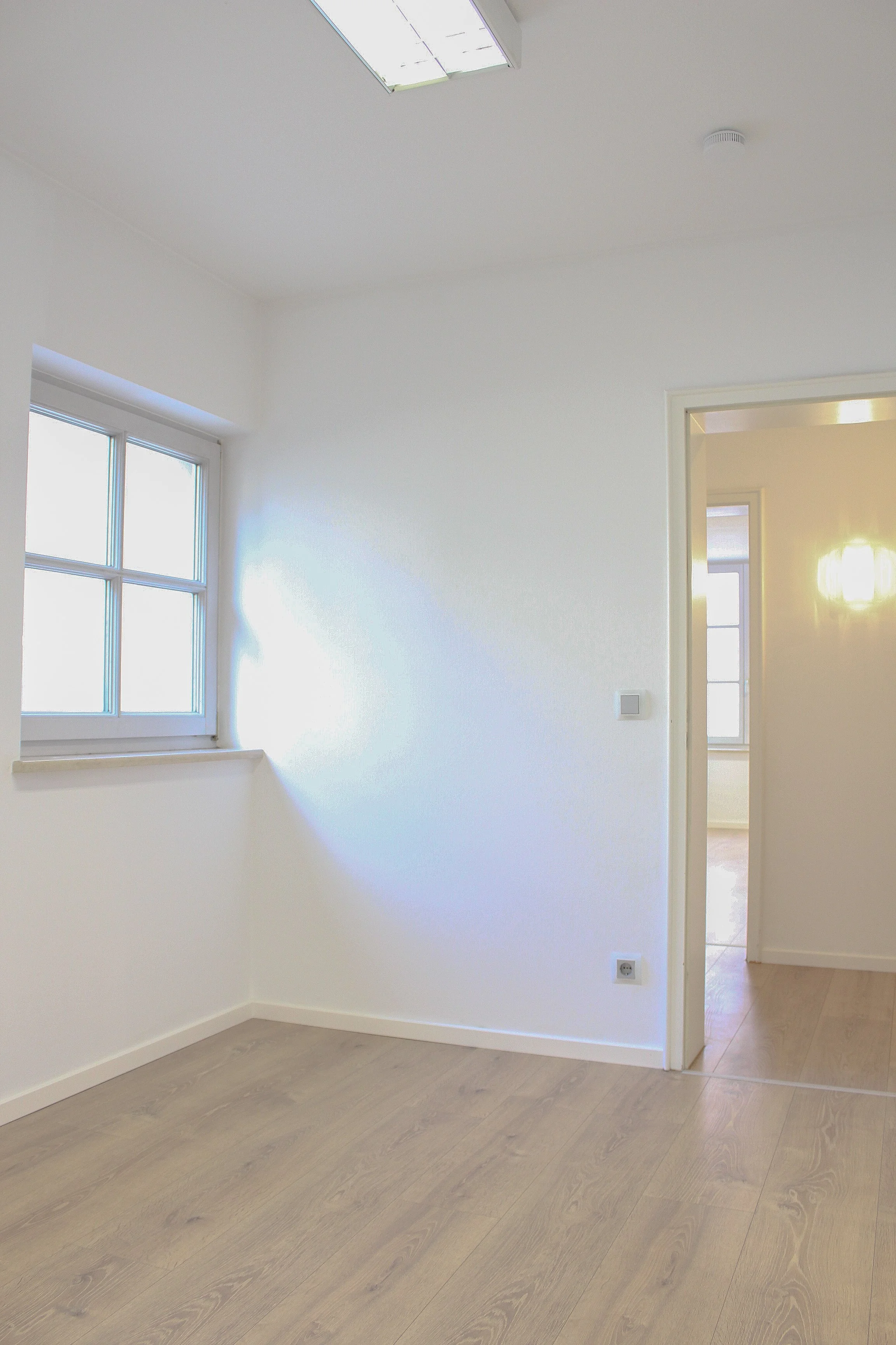 Ein heller, leerer Raum mit weißverputzten Wänden, einer Fensterfront auf der linken Seite, einem offenen Türrahmen auf der rechten Seite und einem Holzboden.