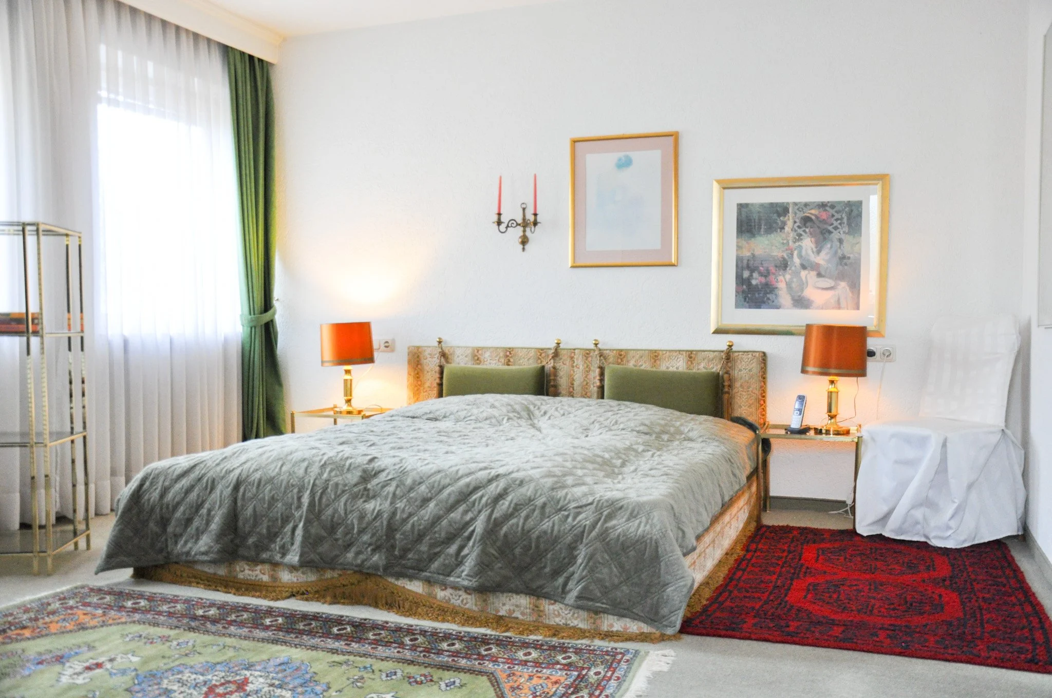 Ein Schlafzimmer mit einem Bett, zwei Lampen, Bilder an der Wand, einem Teppich und Vorhängen am Fenster.