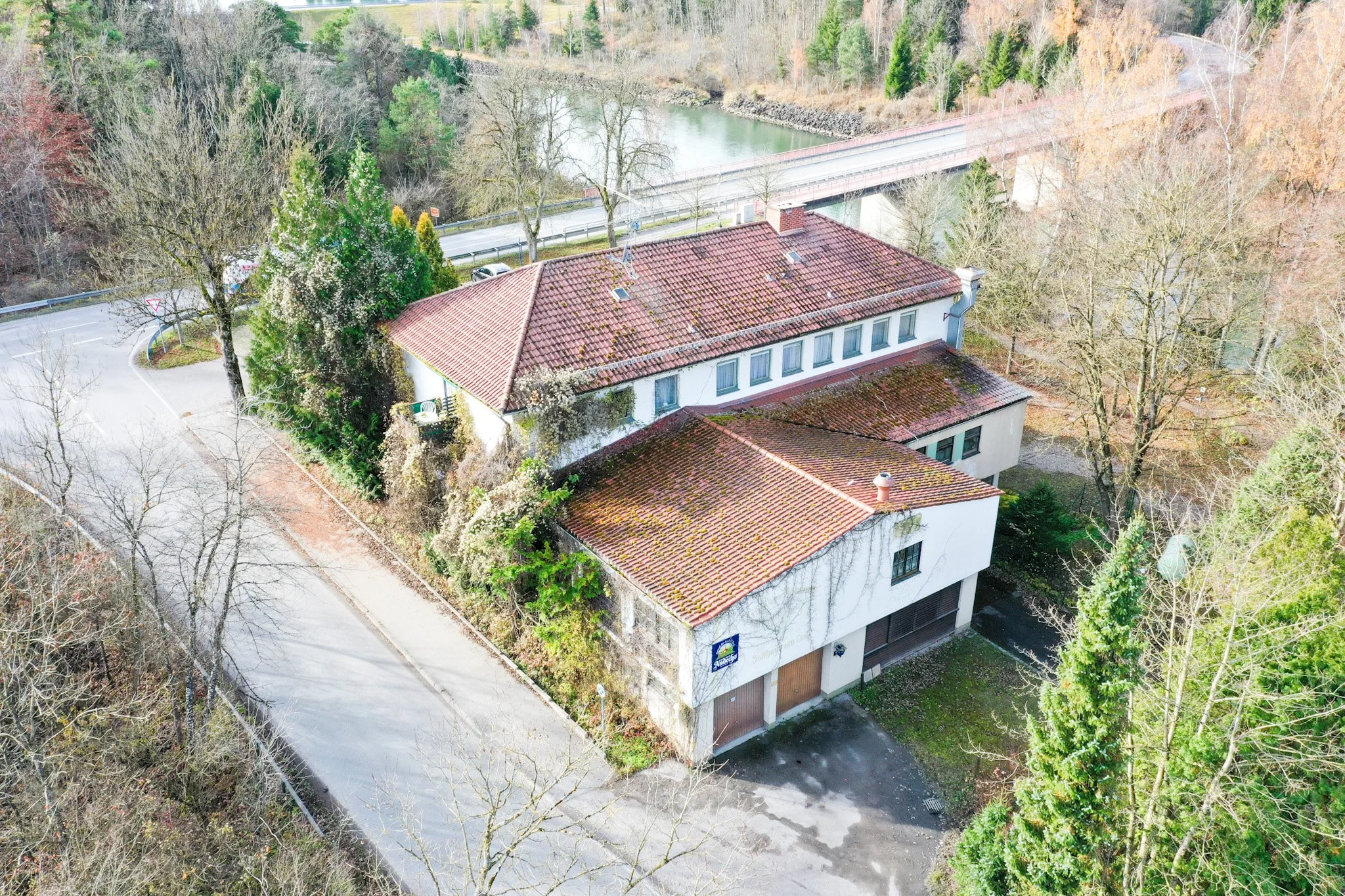 Vogelperspektive auf ein Haus mit rotem Ziegel-Dach, umgeben von Bäumen, an einem Fluss und einer Straße in der Natur gelegen.