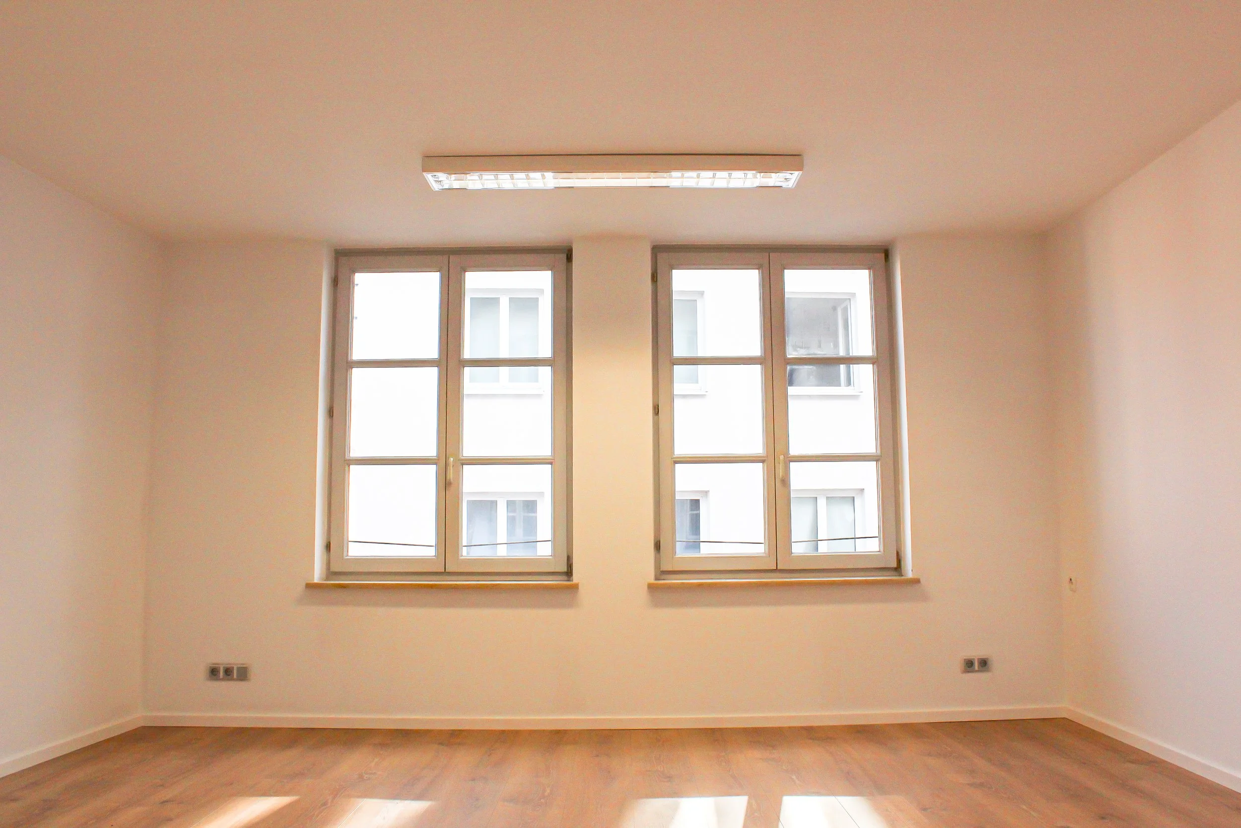 Leeres Zimmer mit großen Fenstern, weißen Wänden und Holzboden.