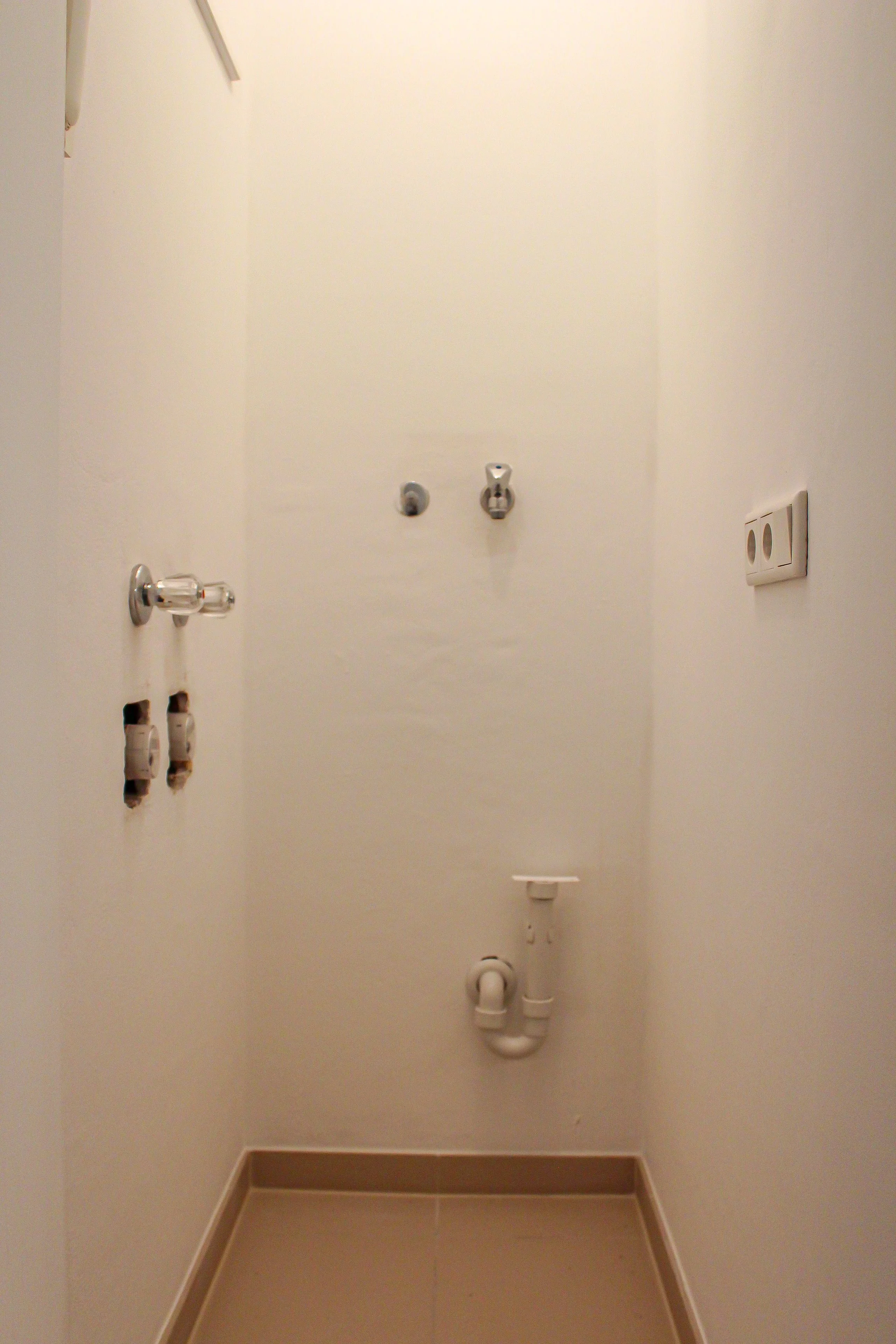 Ein leerer Waschraum mit weißen Wänden, weißen Wasserhähnen, Steckdosen und Abwasserleitungen an der Wand, und einem beigefarbenen Boden.