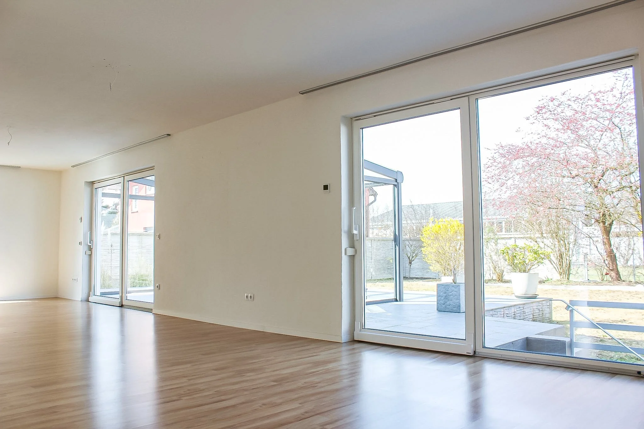 Helles, leeres Zimmer mit großen Fenstern und Holzfußboden, Blick nach draußen auf Bäume und Garten.