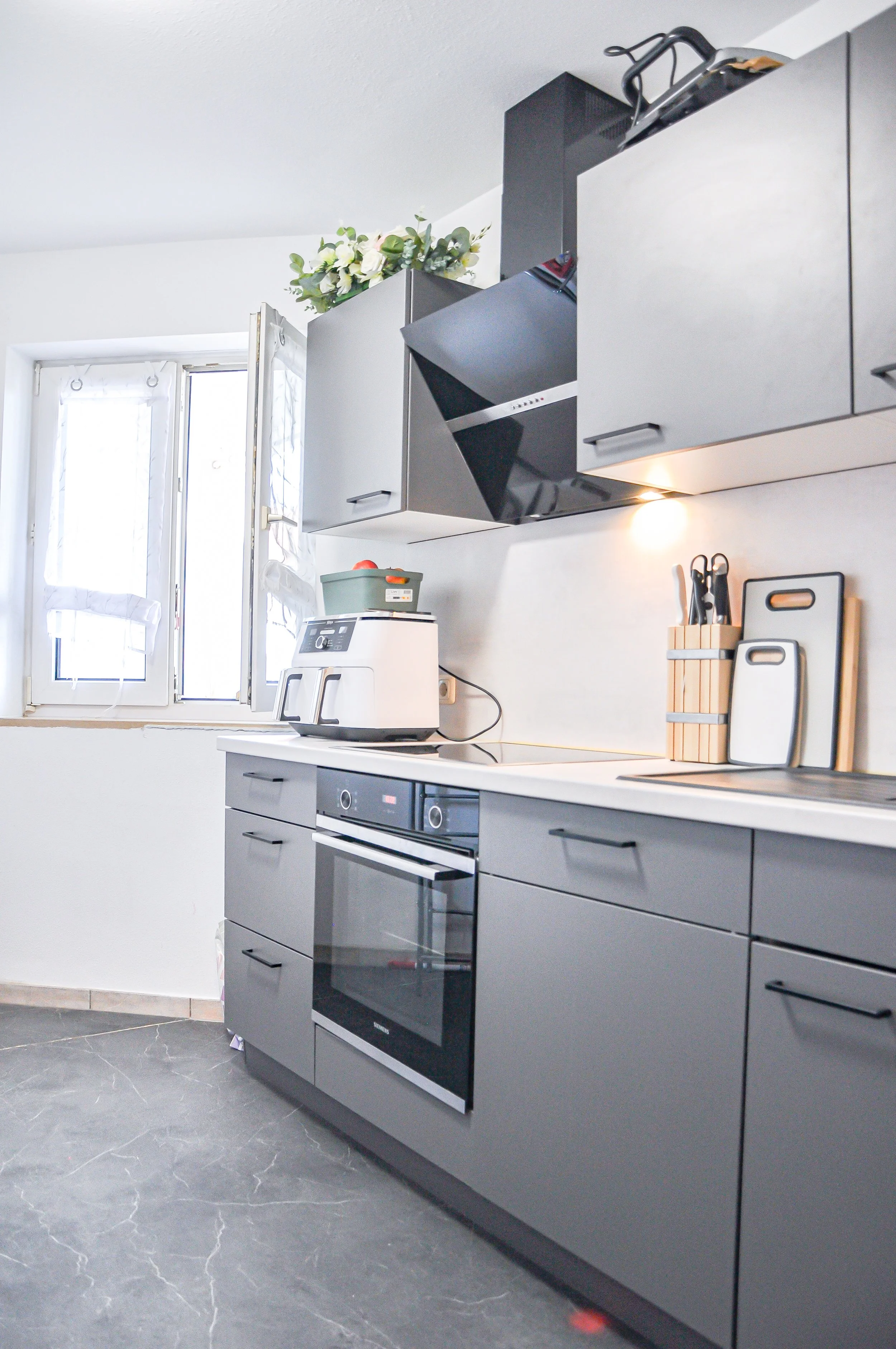 Küche mit grauen Schränken, schwarzer Herd, weißen Wänden, einem Fenster, einem Gasherd mit Backofen, Küchenutensilien im Messerblock, einer Küchenrolle und einem Küchenmesser auf der Arbeitsplatte, und einem Ventilator auf dem Küchenschrank