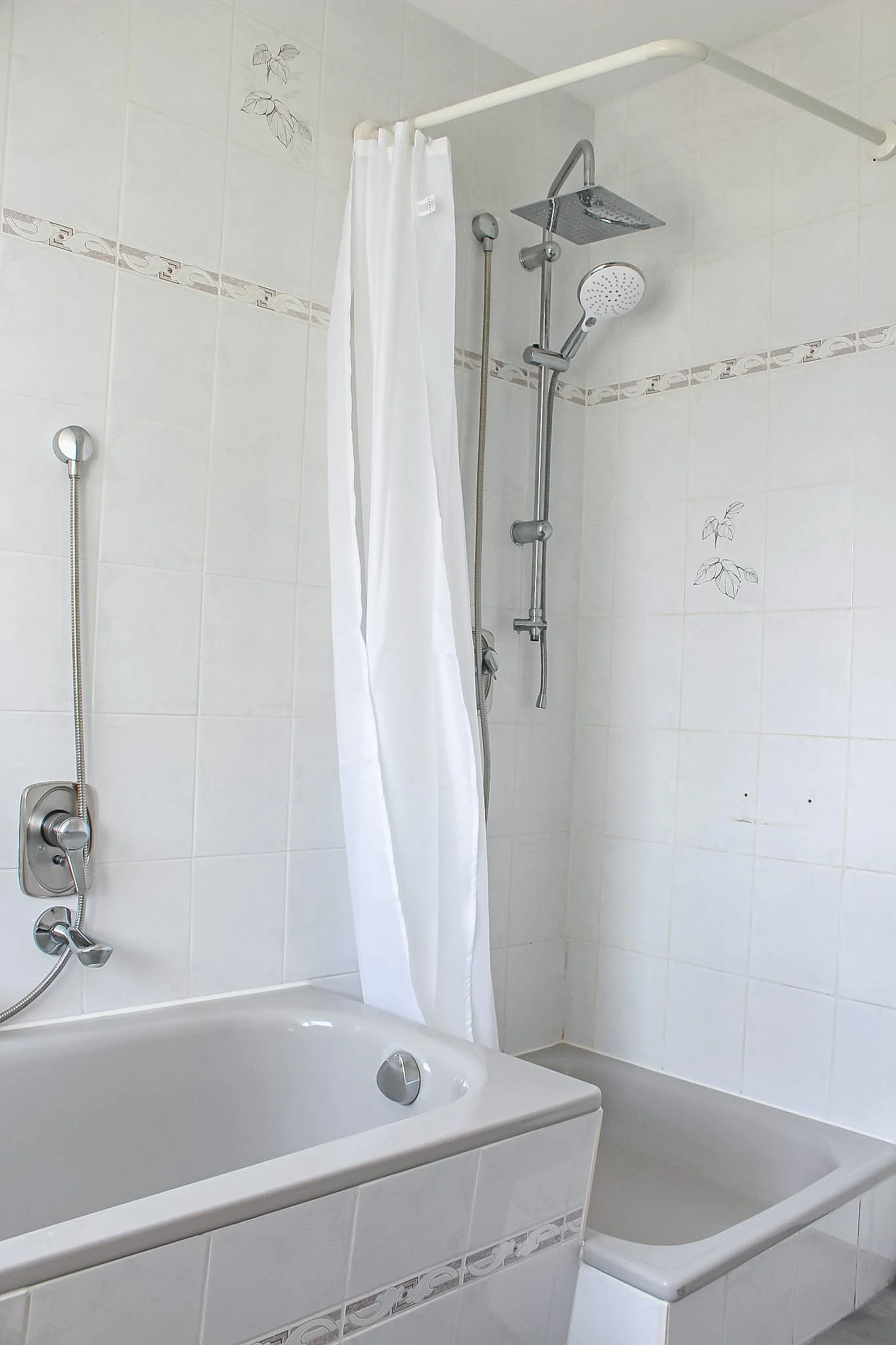 Ein Badezimmer mit Badewanne, Duschkopf, Duschhahn und weißen Fliesen mit einer Pflanzenmuster-Dekoration.