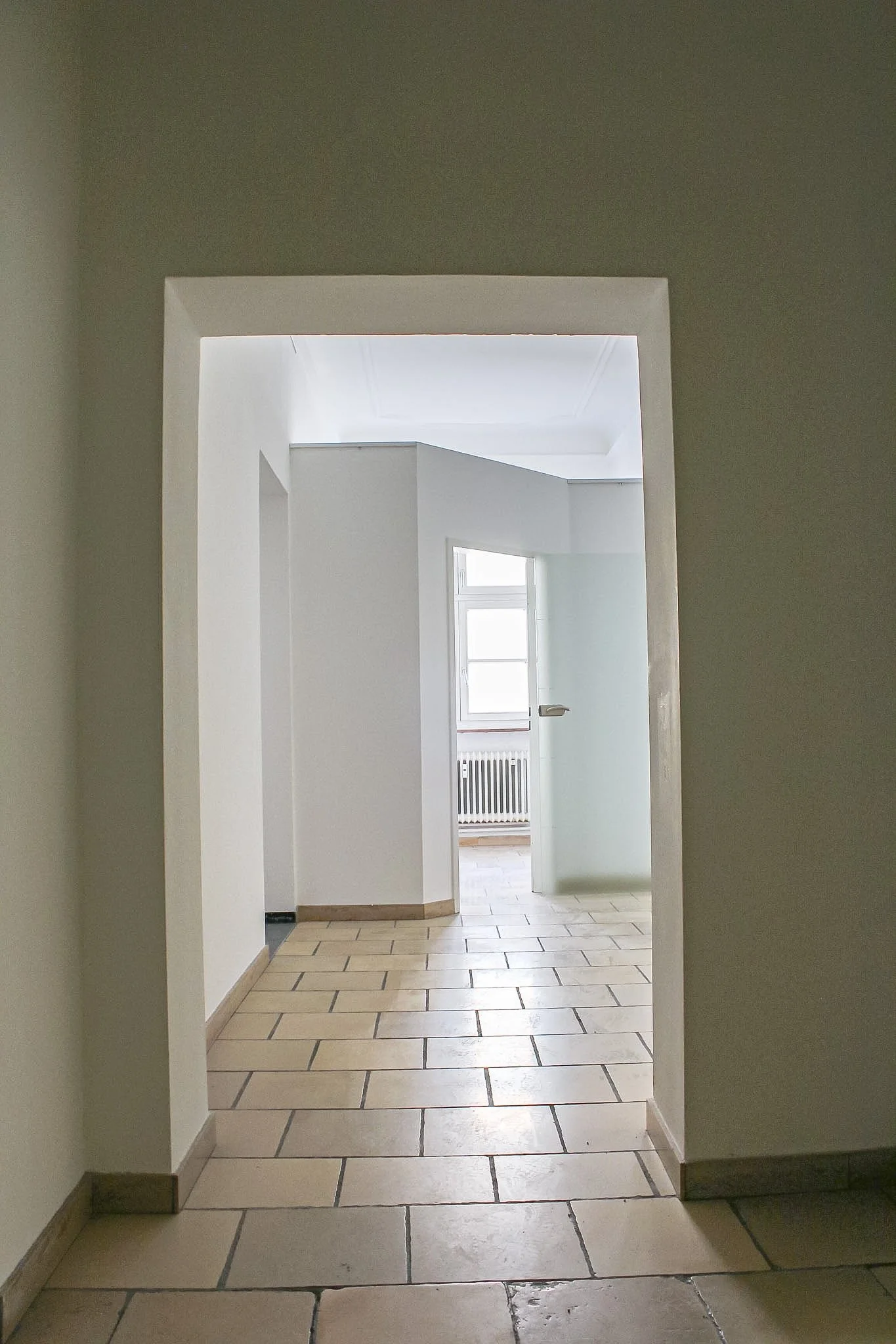 Innenaufnahme eines Flurs mit Fliesenboden und weißen Wänden, Blick durch eine Tür in einen hell erleuchteten Raum mit einem Fenster, Radiator und weißen Wänden.