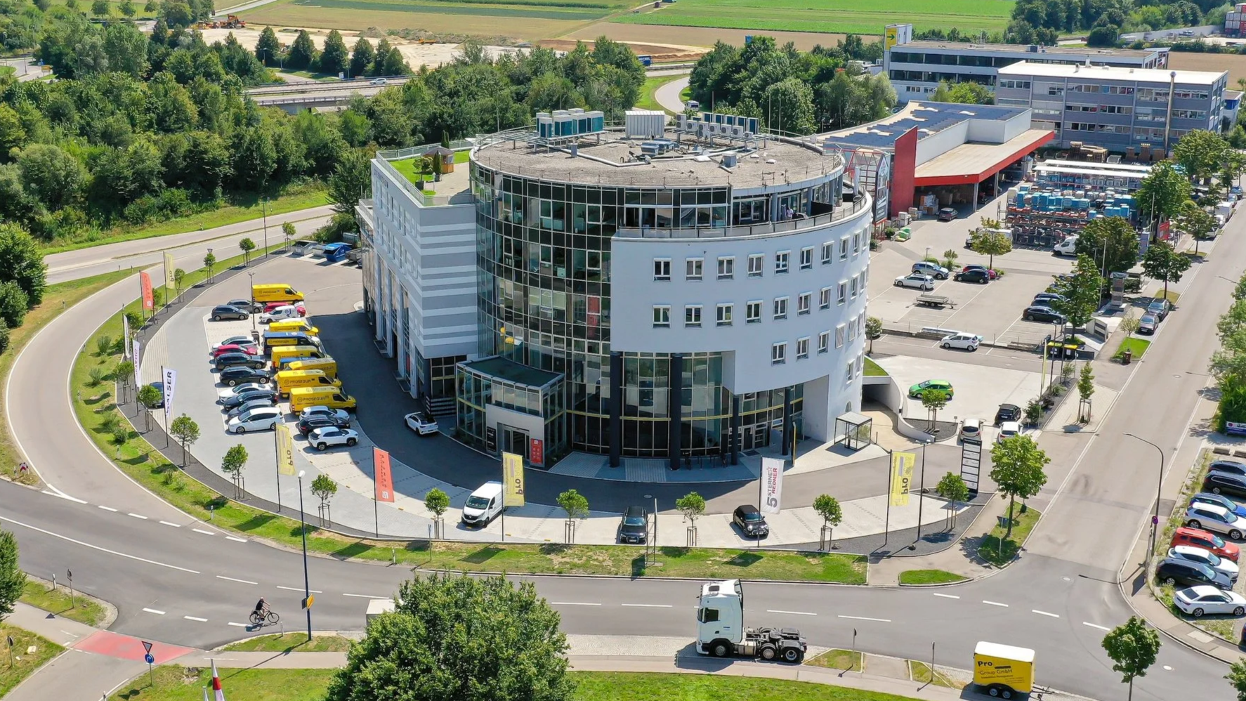 Ein modernes Bürogebäude mit runden und rechteckigen Fassaden, umgeben von Parkplätzen mit Autos, Bäumen und Straßen, im Hintergrund grüne Felder und Wälder.