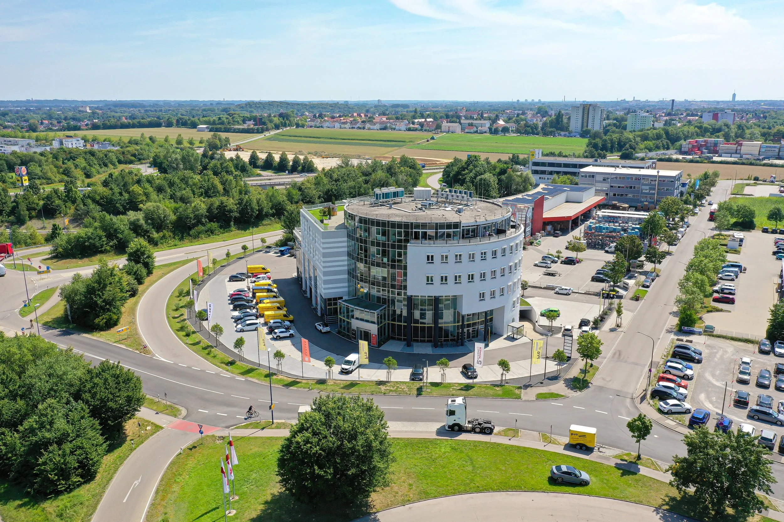 Ein moderner, runder Gebäudekomplex mit vielen Fenstern und einem Parkhaus. Die Umgebung besteht aus Straßen, Bäumen und einem Landstraße mit grünen Feldern im Hintergrund.