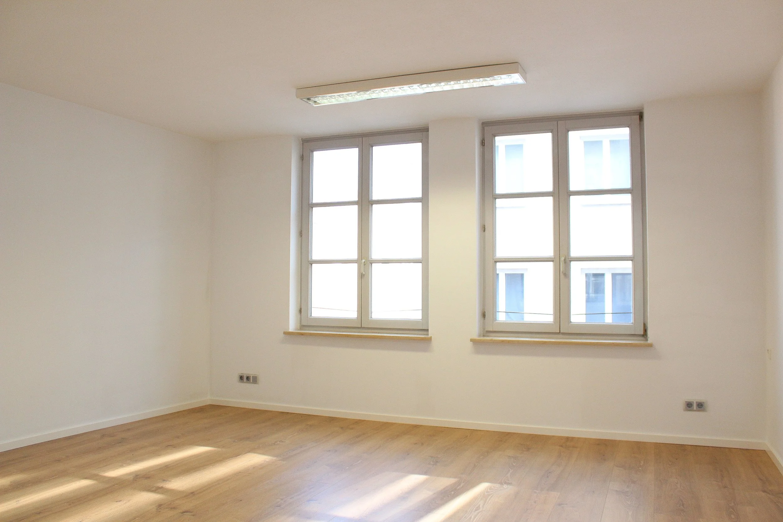 Ein leeres Zimmer mit weißen Wänden, großen Fenstern und Holzboden.