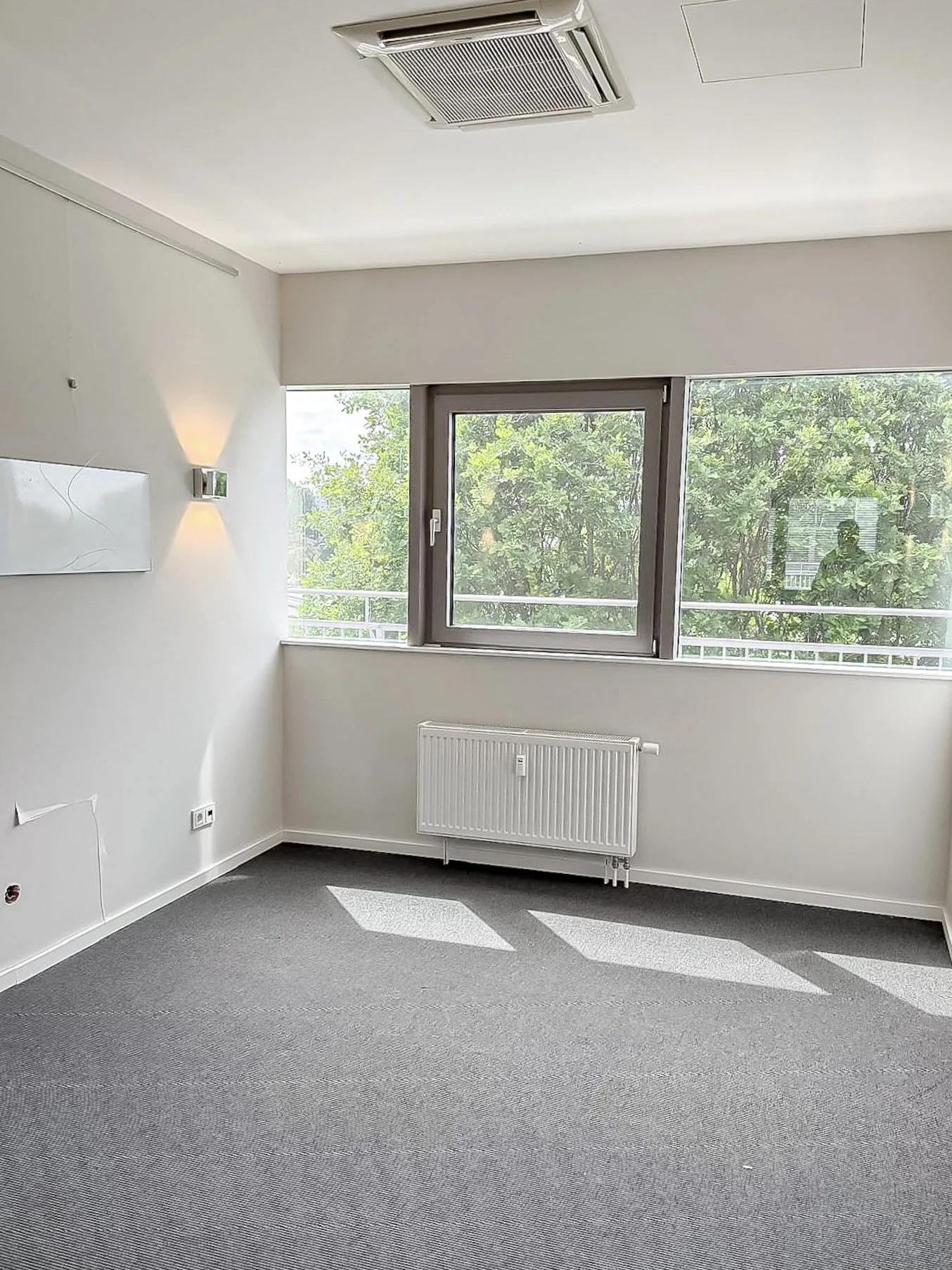 Leerer heller Raum mit grauem Teppichboden, weißen Wänden, einem großen Fenster mit eingeschaltetem Rollladen, Heizkörper darunter, Wandlampen, mehreren Steckdosen und einem Heizregister.