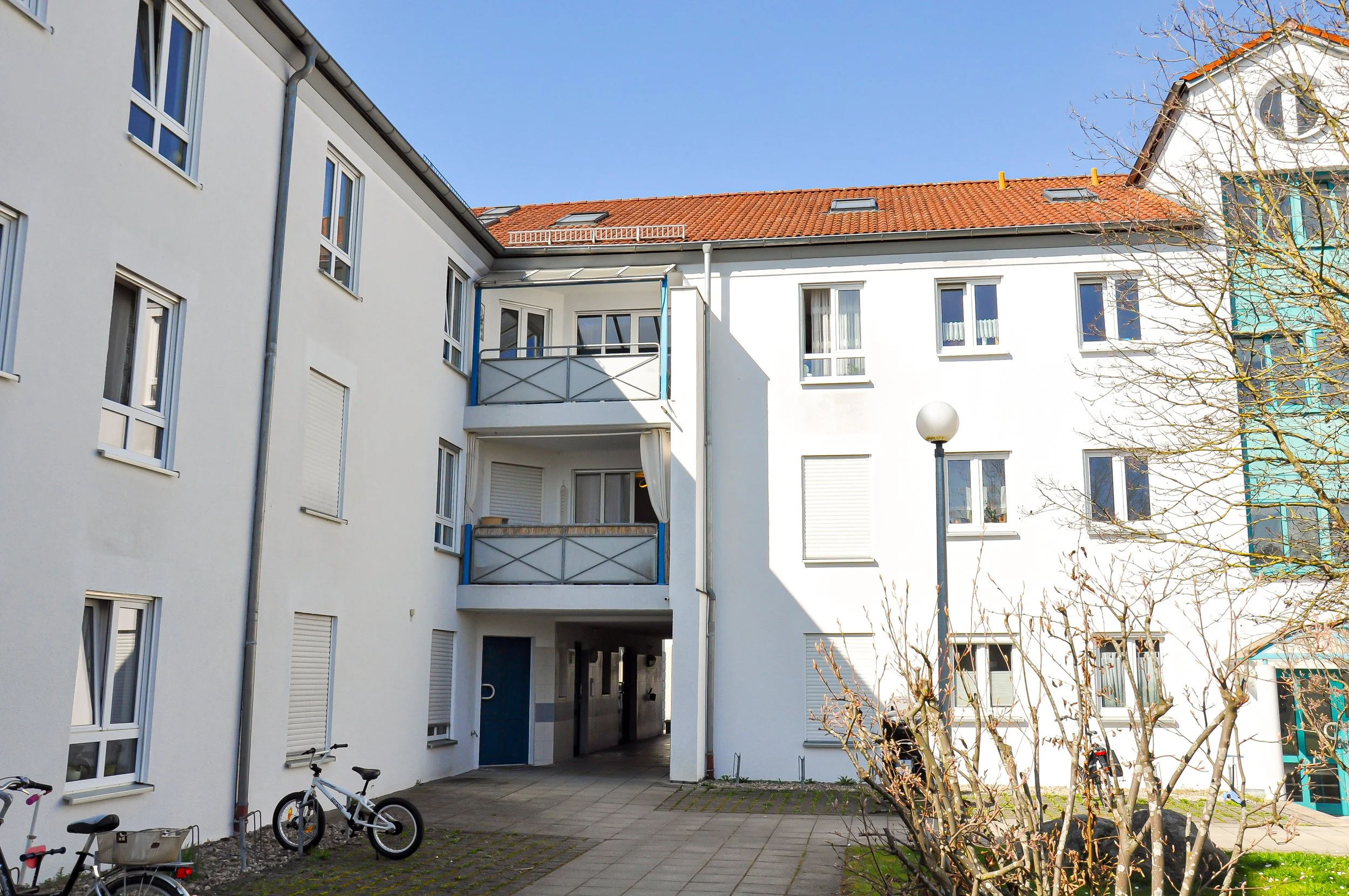 Ein moderner weißer Wohnblock mit mehreren Fenstern und ich Blick auf einen Innenhof, in dem Fahrräder stehen. Der Himmel ist klar und blau, Bäume im Vordergrund haben keine Blätter.
