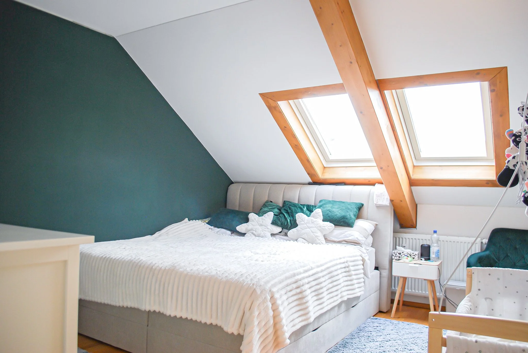 Schlafzimmer mit weißem Bett, grünen Kissen, großen Dachfenstern und Holzbalken, sichtbare Kleider und Puppen auf Nachttisch und Fensterbank, moderner Einrichtungsstil.