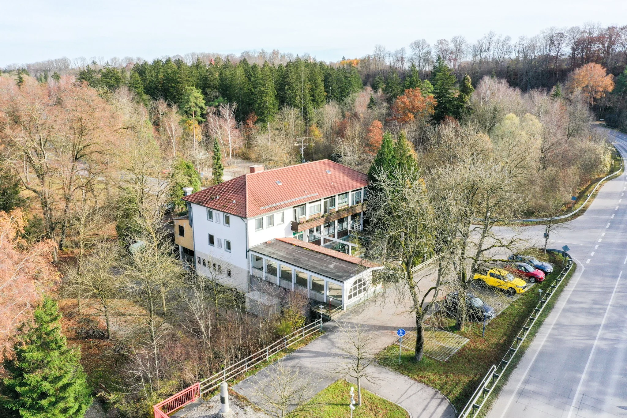 Ein Mehrfamilienhaus mit rotem Ziegeldach umgeben von Bäumen, in einer ruhigen ländlichen Gegend, mit einer Kurve der Straße im Vordergrund und einem Parkplatz mit Autos.