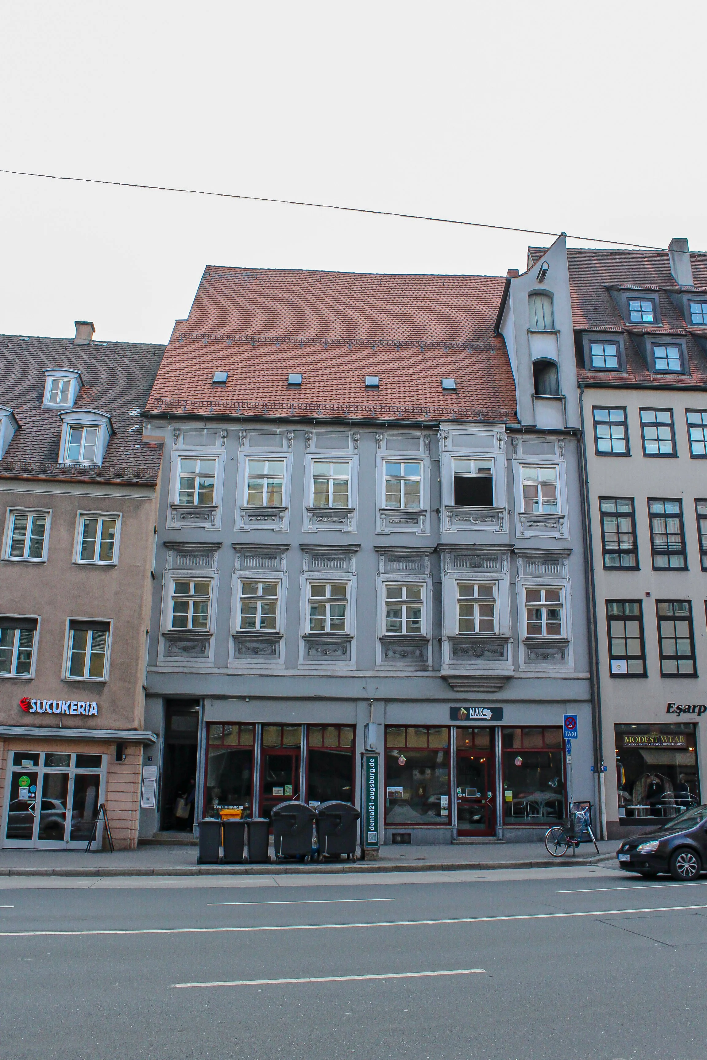 Mehrstöckiges Gebäude mit weißen Fassaden und roten Ziegeldächern, Geschäfte im Erdgeschoss, Fahrrad und Mülltonnen auf dem Bürgersteig, Straßenszene in einer Stadt, auf Deutsch