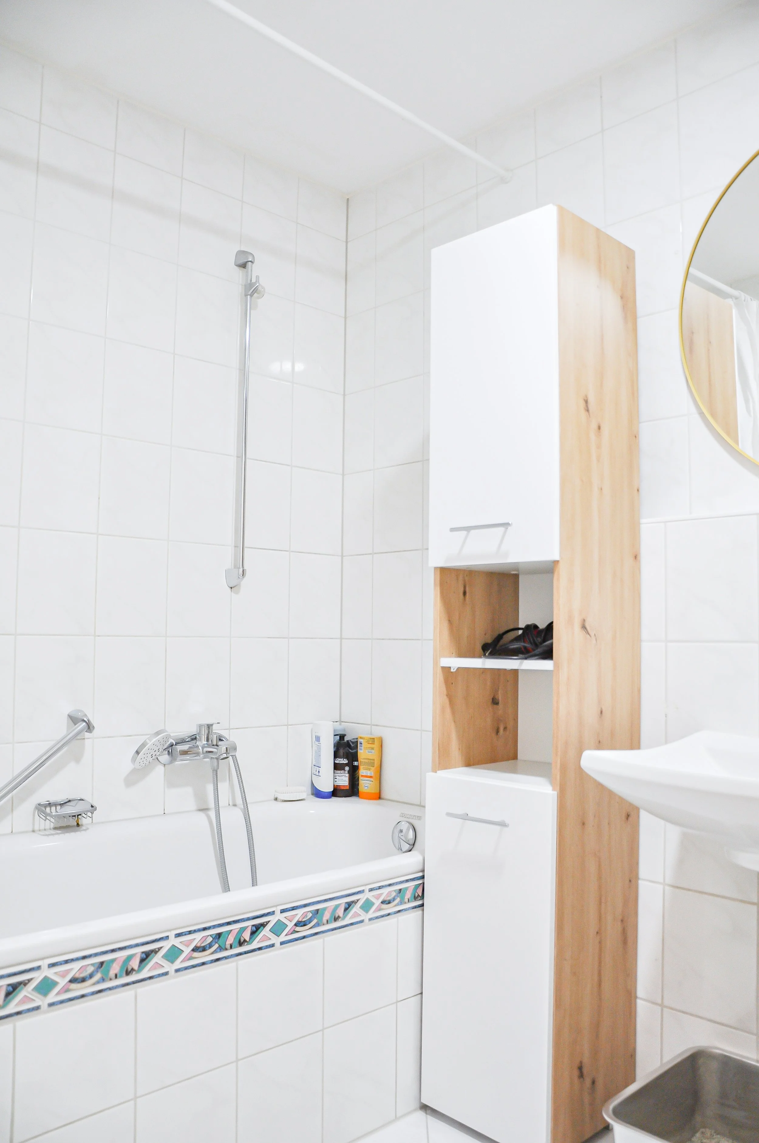 Badezimmer mit Badewanne, Waschbecken, Schränken und verschiedenen Pflegeprodukten, gekachelt mit weißer Wandfliesen, kleine bunte Flieseneinfassung an der Badewanne