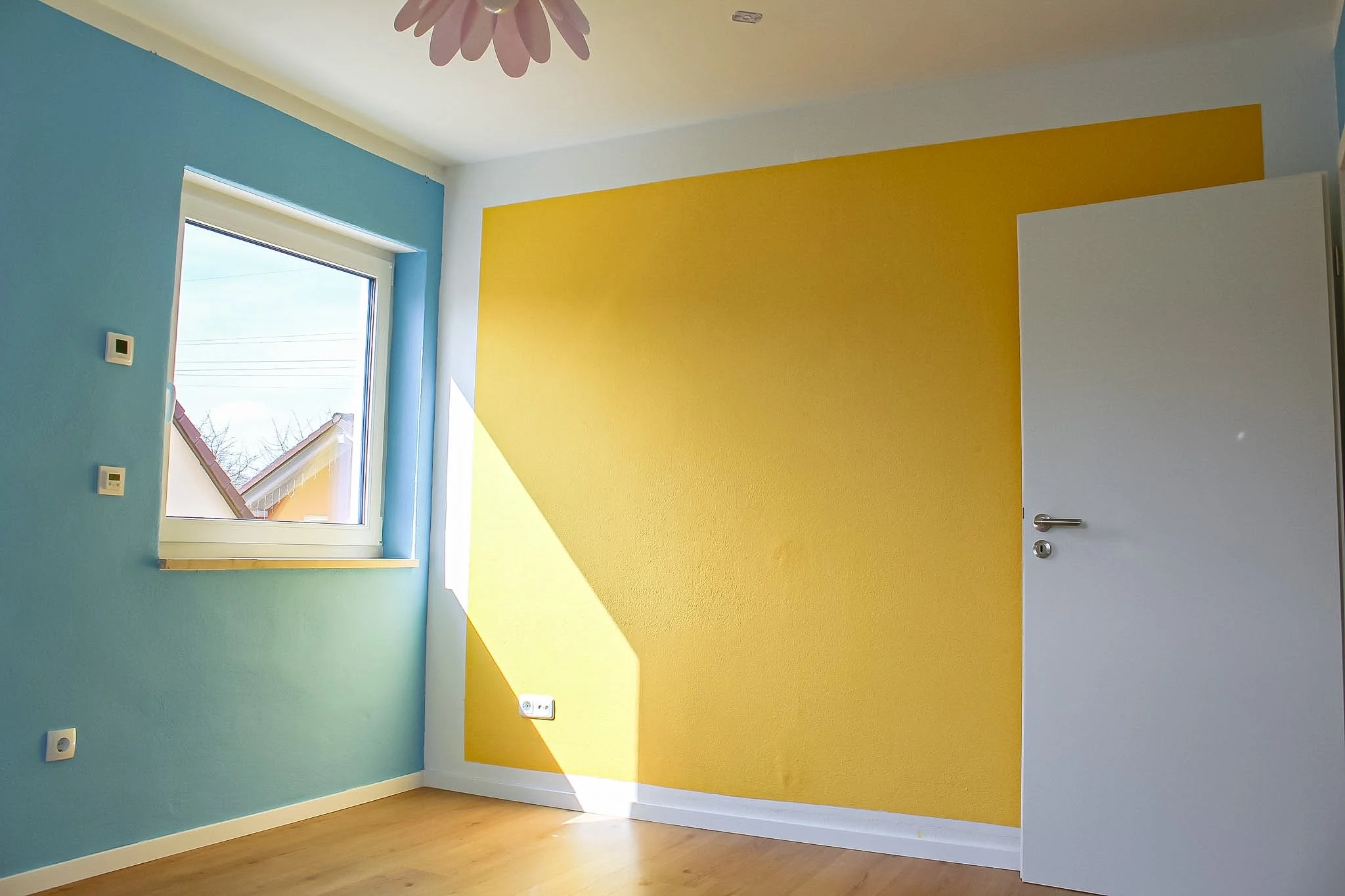 Ein Raum mit blauer und gelber Wandfarbe, ein offenes weißes Tür, ein Fenster mit natürlichem Licht, ein blumiger Kronleuchter, Holzwohnzimmerboden.