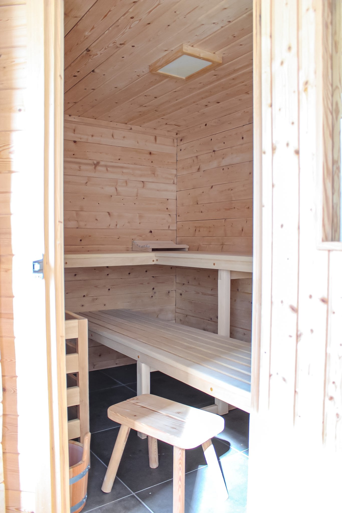 Innenraum eines Holzsaunaraums mit einer Holzbank, einem kleinen Tisch und einem Holzofen auf Fliesenboden.