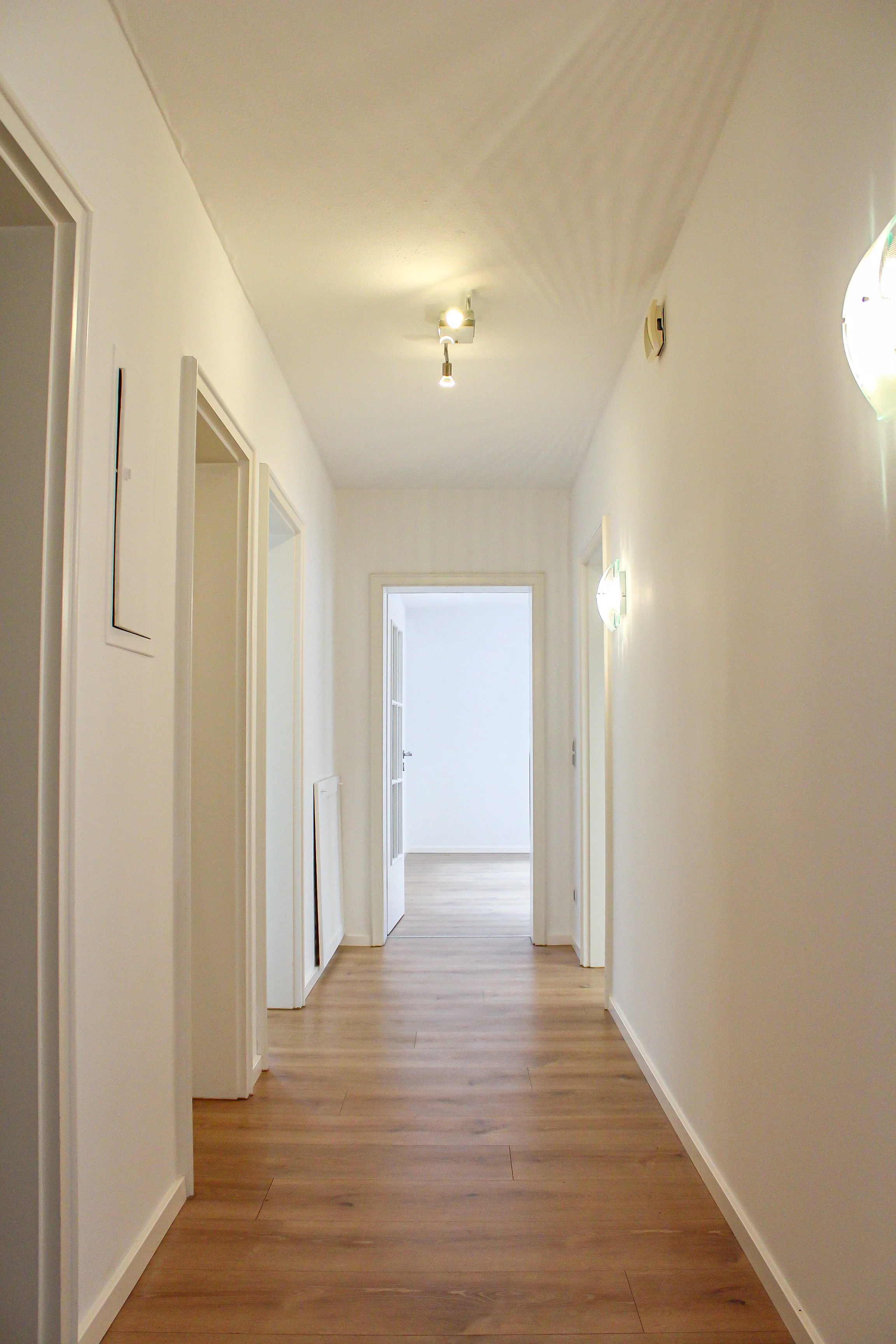 Heller Flur mit Holzfußboden, weiß gestrichene Wände und mehreren verschlossenen Türen auf beiden Seiten, leuchterbeleuchtung an der Decke und Wandlampen, im Hintergrund eine offene Tür zu einem hell erleuchteten Raum.
