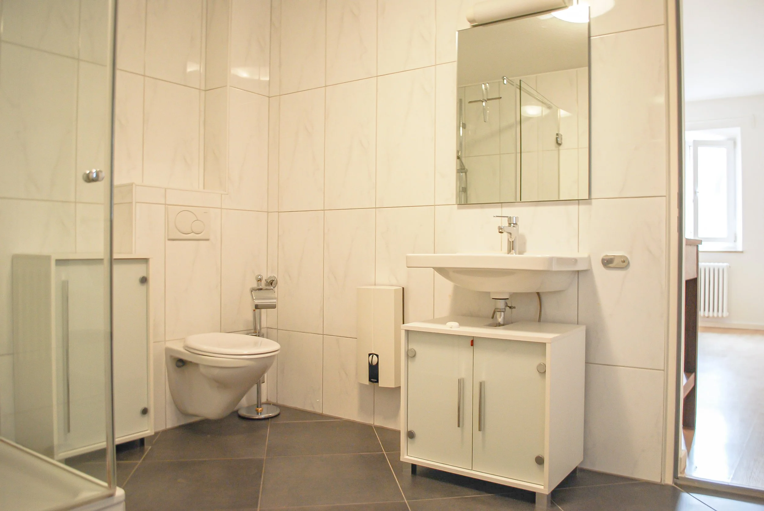 Kleine Badezimmer mit weißen Fliesen, Toilette, Spiegel, Waschbecken und einem kleinen Waschtisch, Fenster im Hintergrund.