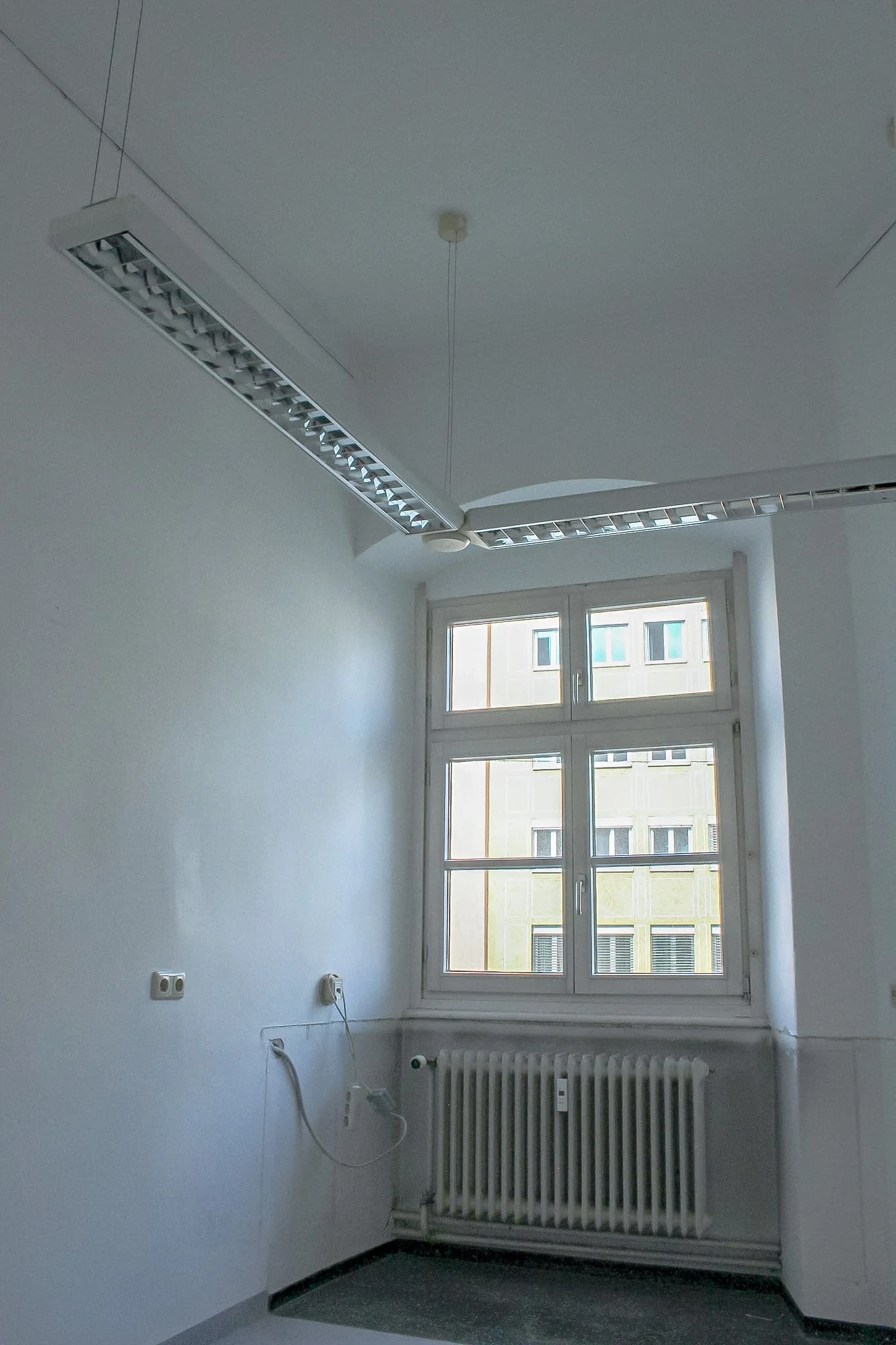 Leerer Büroraum mit weißen Wänden, mehreren Stromanschlüssen, einem Heizkörper, einem Fenster und einer Deckenleuchte.