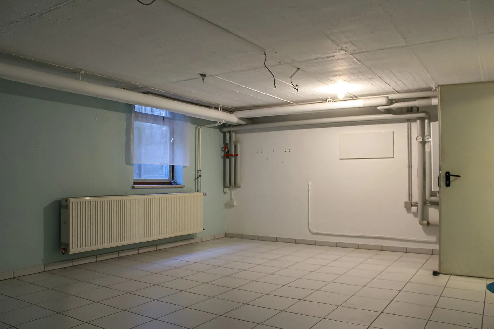 Ein unvollendeter Kellerraum mit weißen Wänden und Fliesen auf dem Boden. In der Ecke sind Wasserleitungen, eine Heizung an der Wand, und das Decken sind Kabelvergewindungen sichtbar.