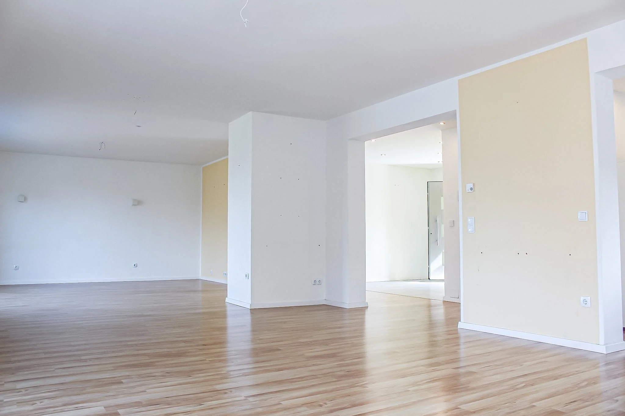 Leeres, gut beleuchtetes Wohnzimmer mit weißen Wänden, Holzboden und offenen Bereichen.