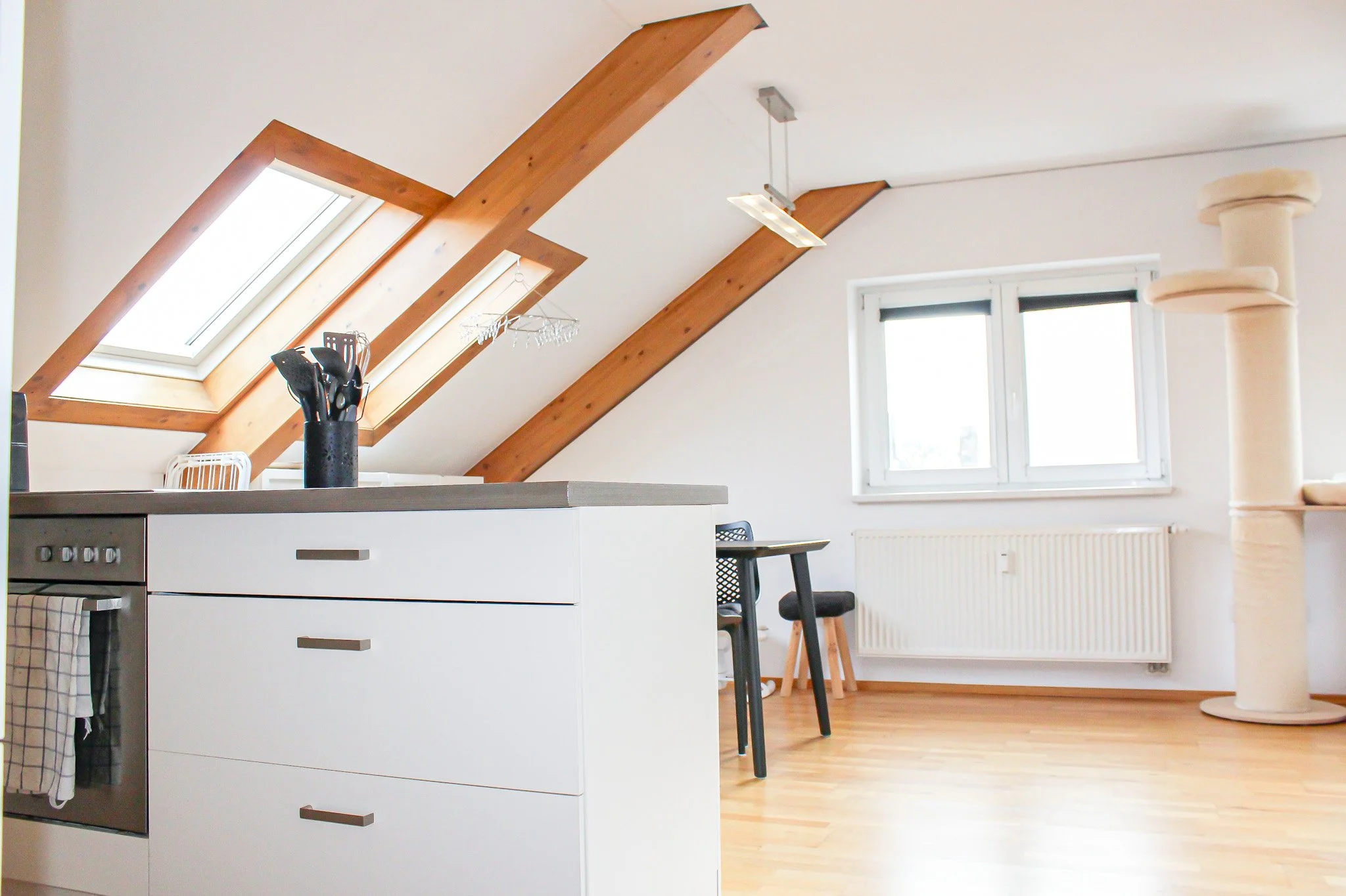 Ein moderner Küchenbereich mit maximaler Nutzung des Dachgeschosses, inklusive Kochinsel, Essbereich, Wandschränken, Fenster und Dachgauben mit Licht. Skandinavischer Stil mit hellem Holz, Weiß und natürlichen Materialien.