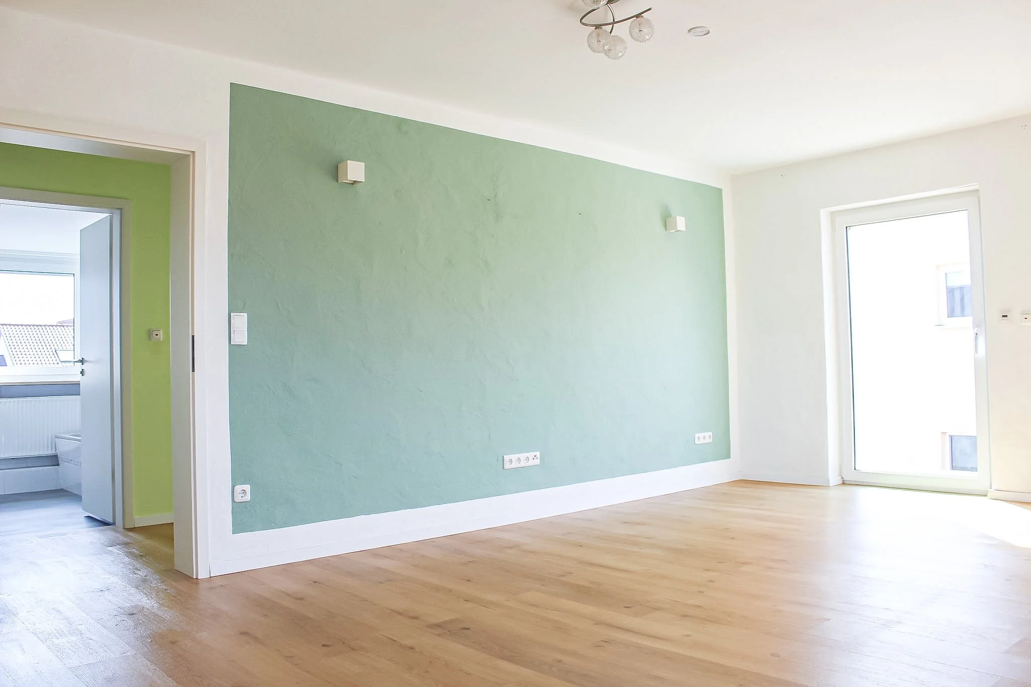 Leeres Wohnzimmer mit grüner Akzentwand, großen Fenstern und hellem Holzparkett.