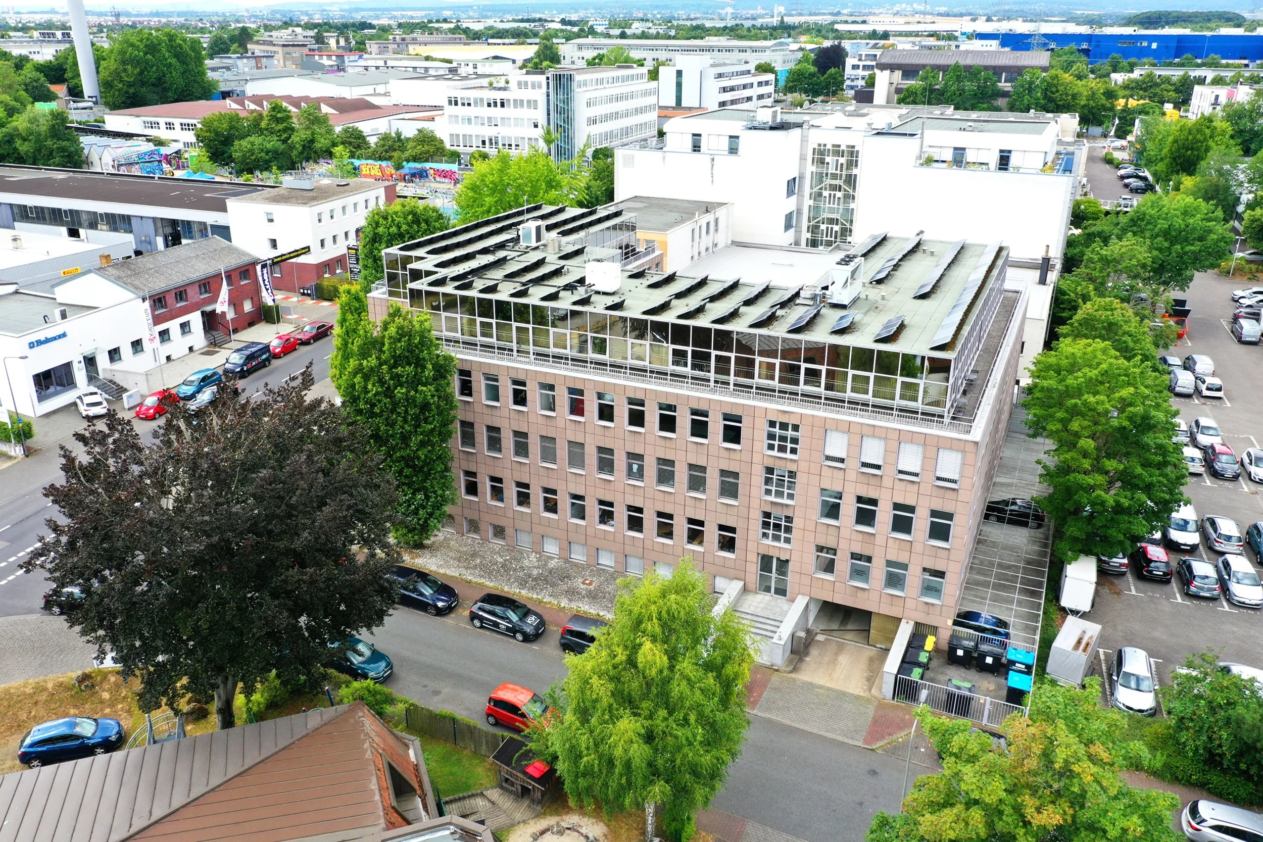 Ein städtischer Blick auf mehrere Bürogebäude und ein Wohnhaus, umgeben von Bäumen und geparkten Autos. Im Vordergrund sind die Dächer eines Gebäudes mit Solarpanelen sichtbar.