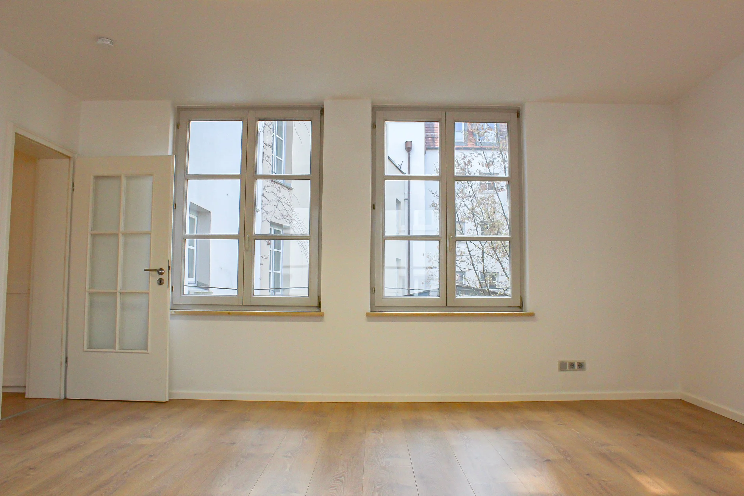 Leeres Zimmer mit weißen Wänden, großen Fenstern und Holzboden.