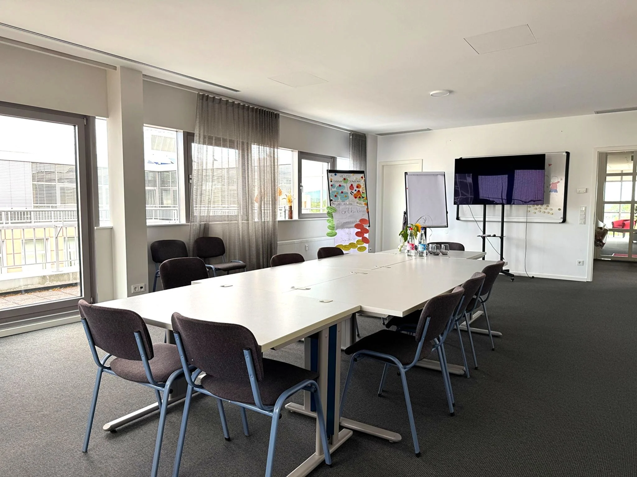 Ein Sitzungsraum mit einem langen weißen Tisch, mehreren schwarzen Stühlen, großen Fenstern mit Vorhängen, einem großen Bildschirm, Whiteboards, Pflanzen und Dekorationen.