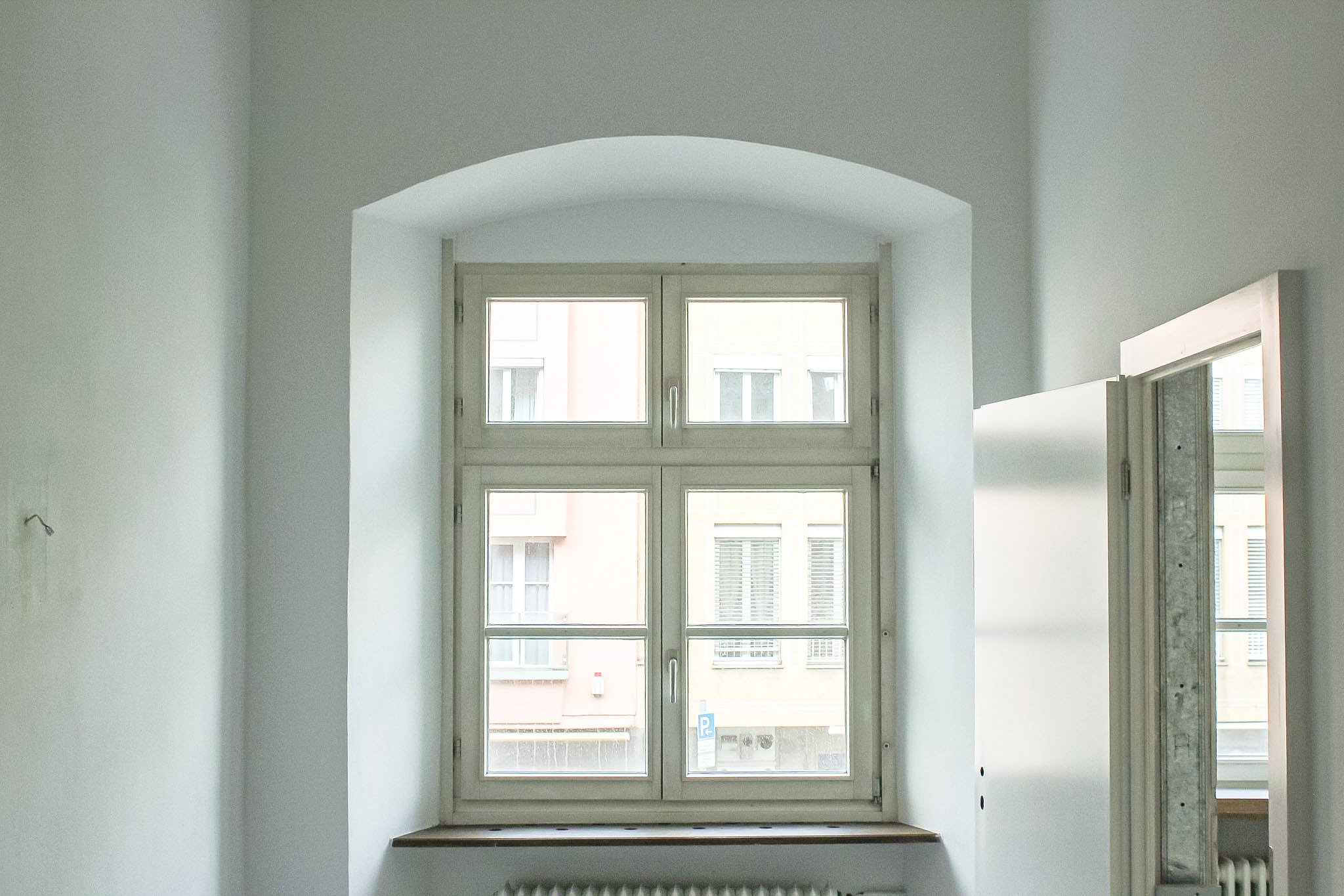 Ein Fenster in einem hellen Raum mit weißen Wänden und einer schwarzen Fensterbank, durch das ein Blick auf Nachbargebäude gegeben ist.