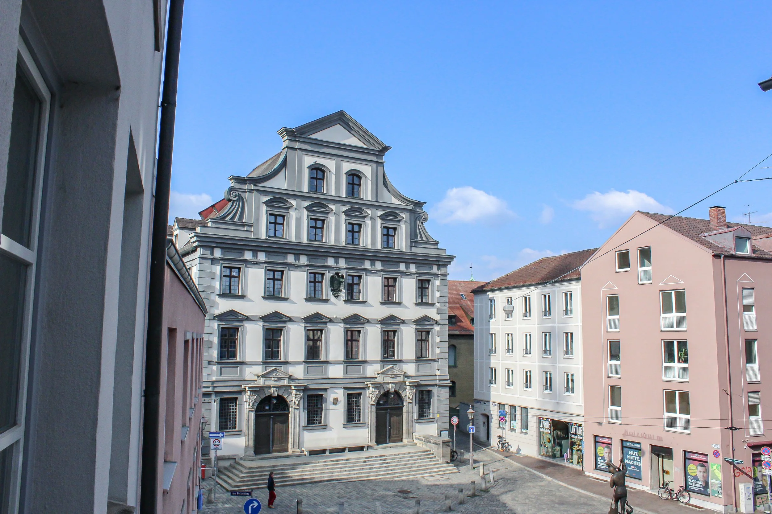 Blick auf eine historische Stadthalle mit barocker Fassade in einer Innenstadt, umgeben von modernen Gebäuden, vor einem blauen Himmel mit wenigen Wolken.