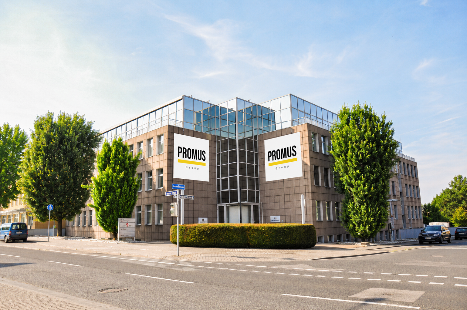 Ein Bürogebäude mit zwei großen Werbetafeln auf der Fassade, umgeben von Bäumen, Straßen und parkenden Autos an einem sonnigen Tag.