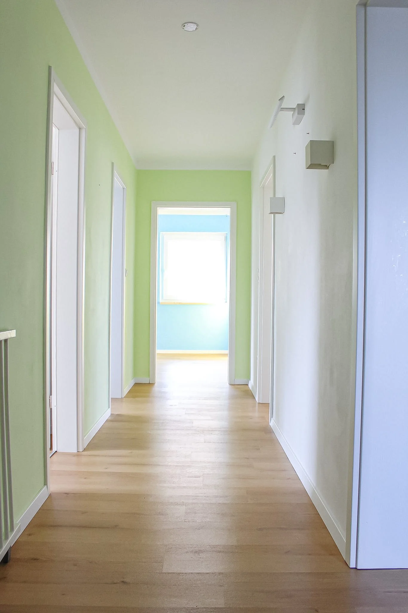 Ein heller Flur in einem Haus, mit grünen Wänden auf der linken Seite und weißen Wänden auf der rechten Seite. Es gibt mehrere Türen an den Seiten und ein Fenster am Ende des Flurs, das natürliches Licht hereinlässt. Der Boden ist aus hellem Holz.
