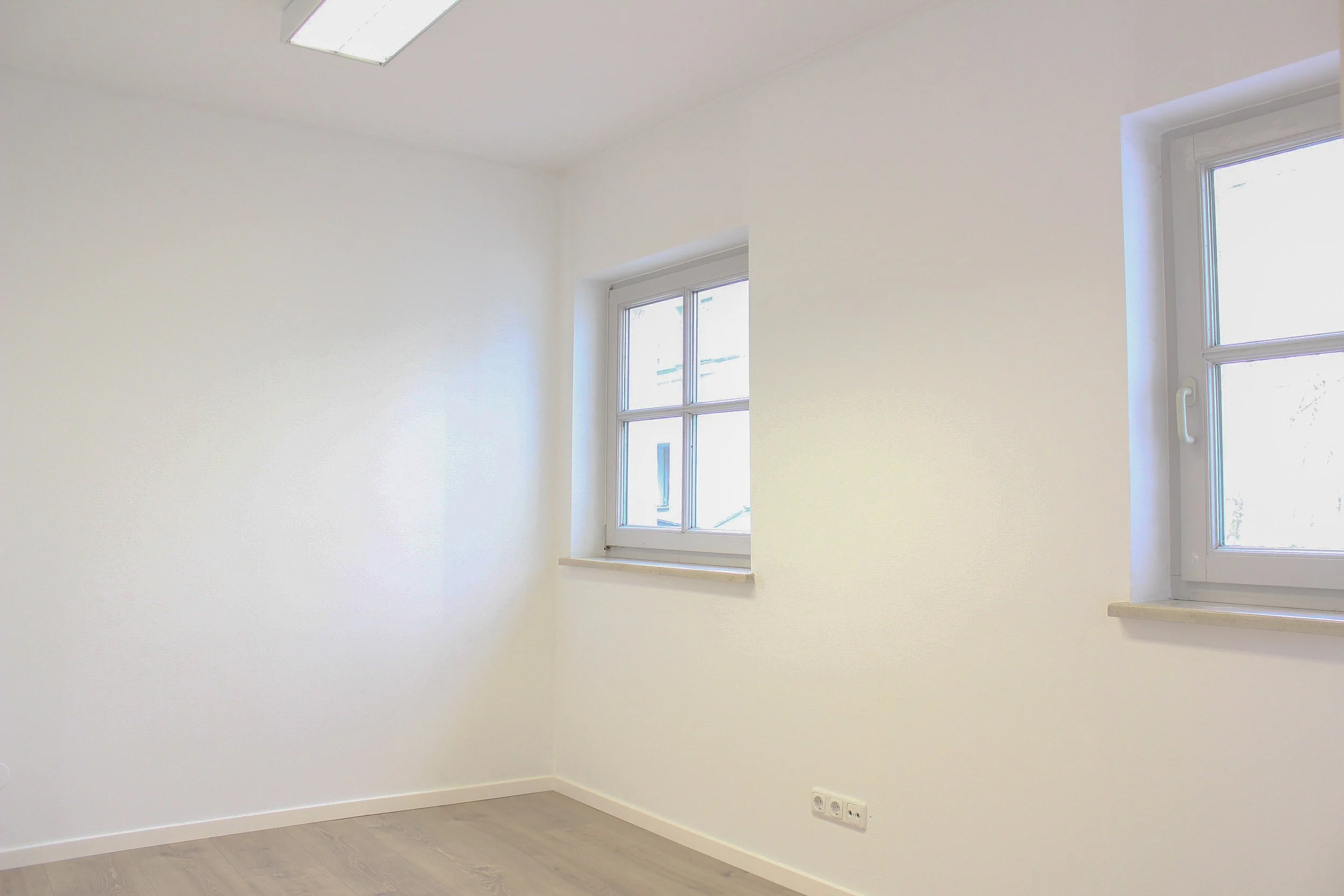 Leeres Raum mit weißen Wänden, zwei Fenster, grauem Boden und Deckenlampen