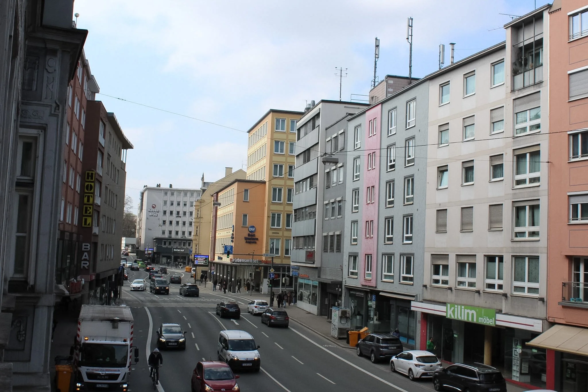 Straße in einer Stadt mit mehreren mehrstöckigen Gebäuden, Autos und Passanten, einige Geschäfte und Schilder an den Gebäuden